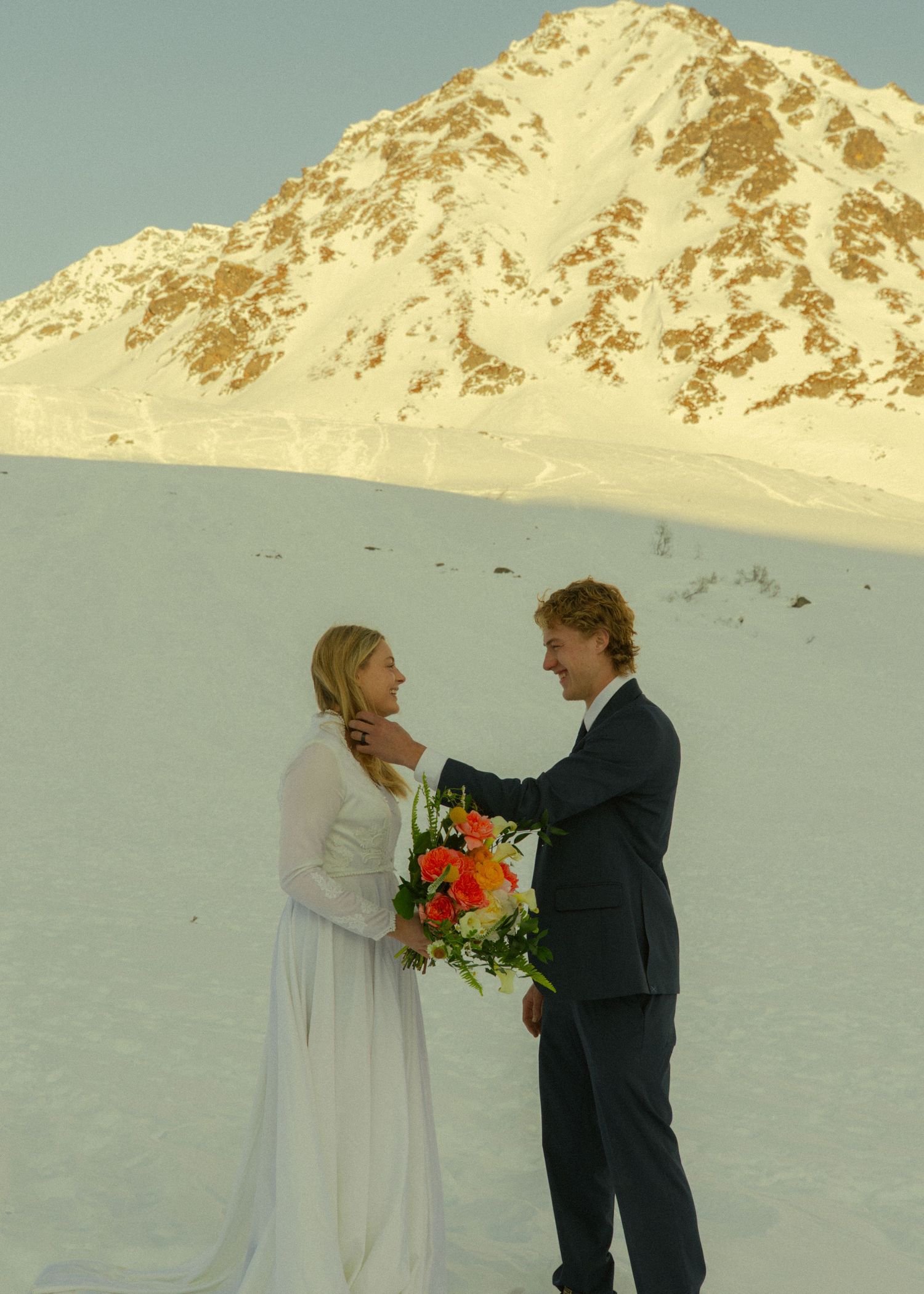 Groom in navy suit tenderly touches bride's face while she holds a vibrant bouquet before a sunlit snowy mountain.