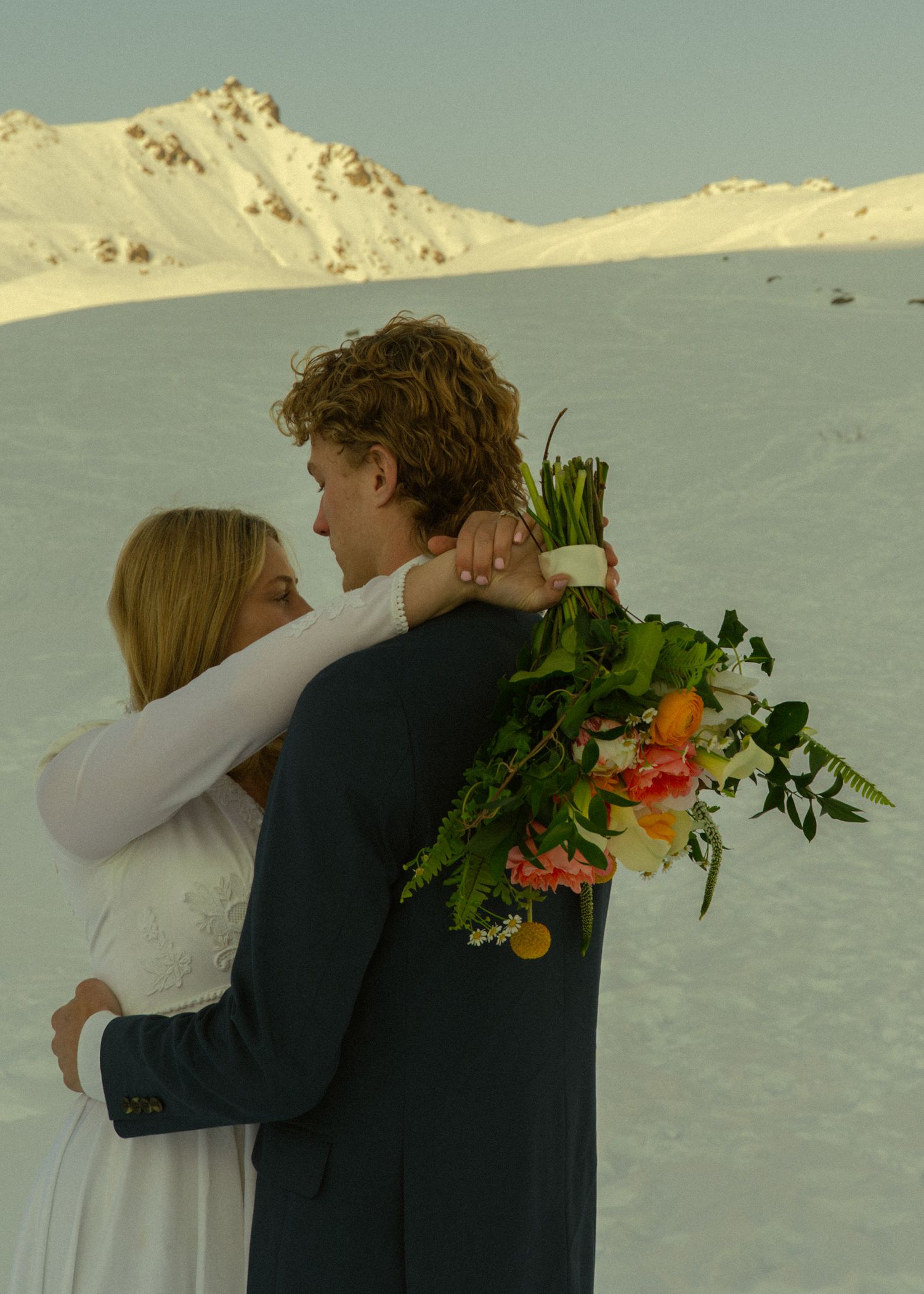 Wedding couple sharing romantic embrace on snowy mountain slope, groom holding vibrant tropical bouquet.