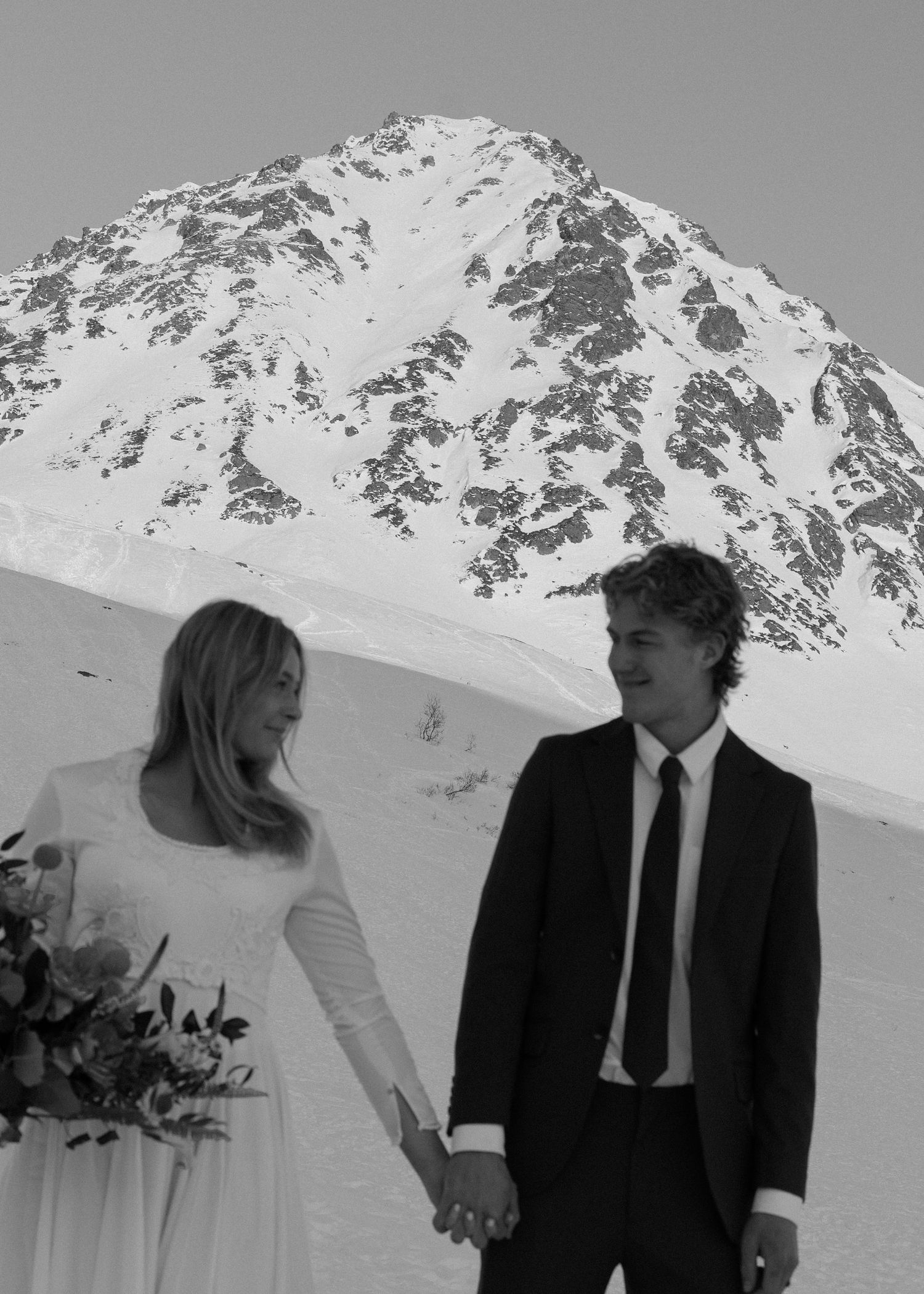 Black and white photo of bride and groom holding hands with a dramatic snow-capped mountain peak behind them.