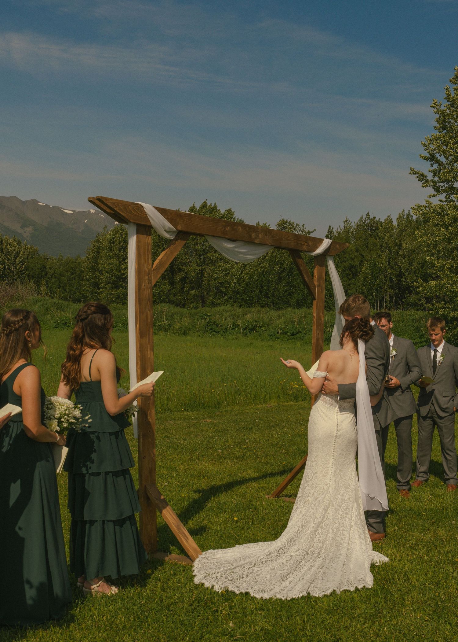Outdoor summer wedding ceremony with bride in lace gown at wooden arch, bridesmaids in forest green dresses.