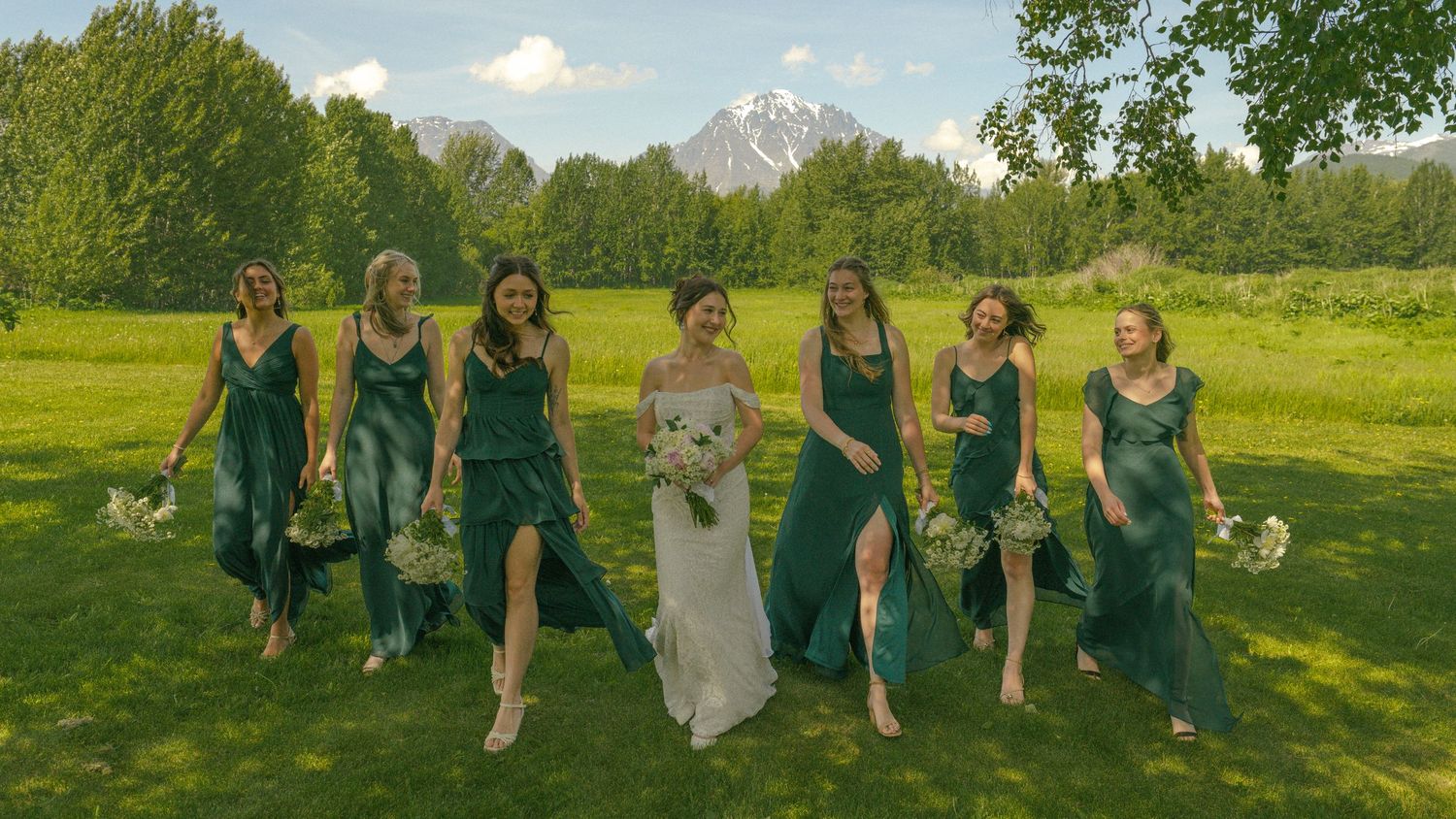 Bride in white gown walks with six bridesmaids in emerald green dresses across sunny mountain meadow.