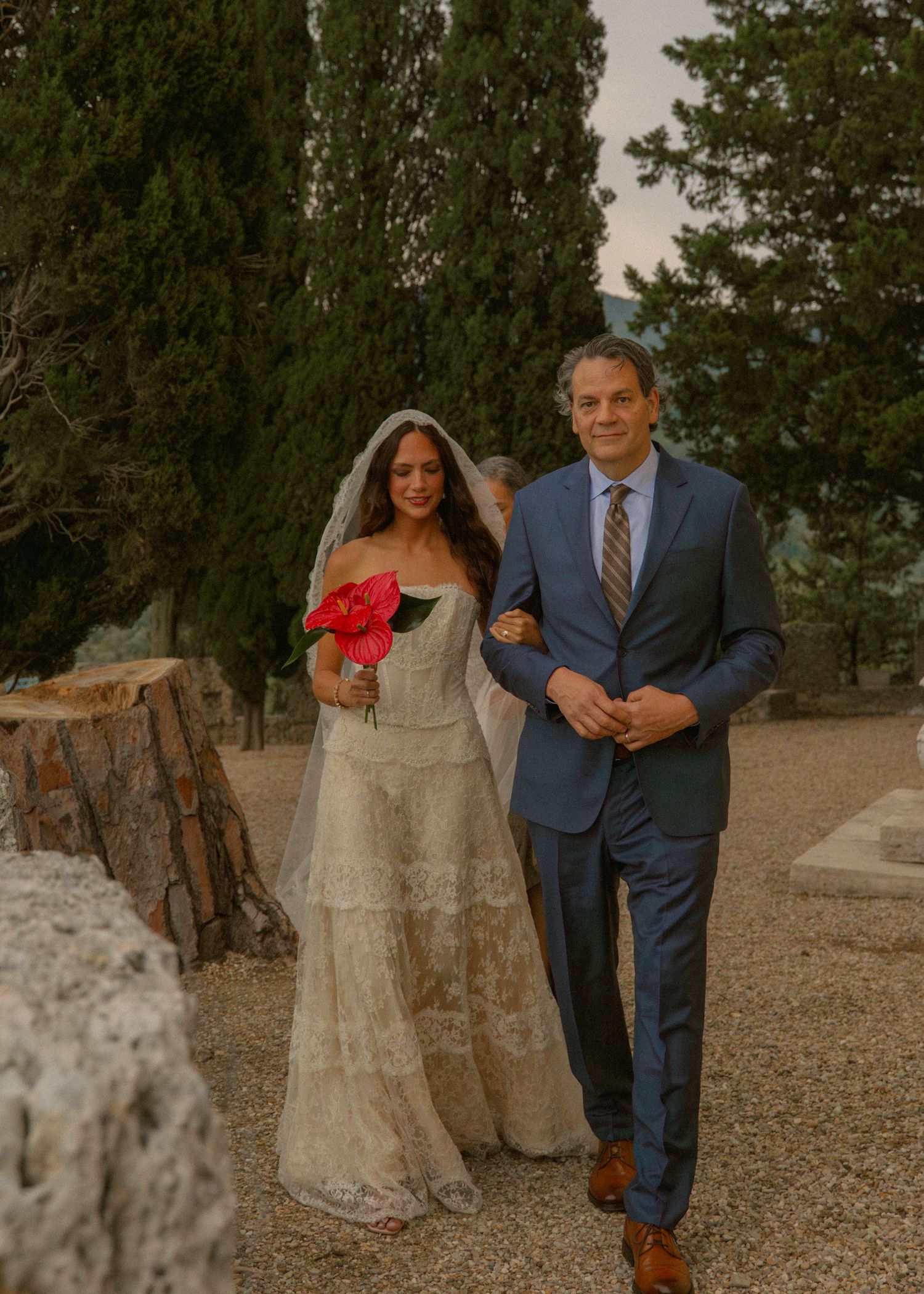 Bride in lace gown holding red rose walks with escort in blue suit at outdoor wedding ceremony.