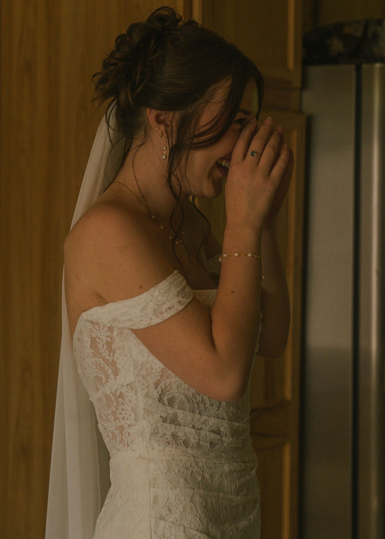 Emotional bride in off-shoulder lace wedding gown and veil covering her face with hands indoors.