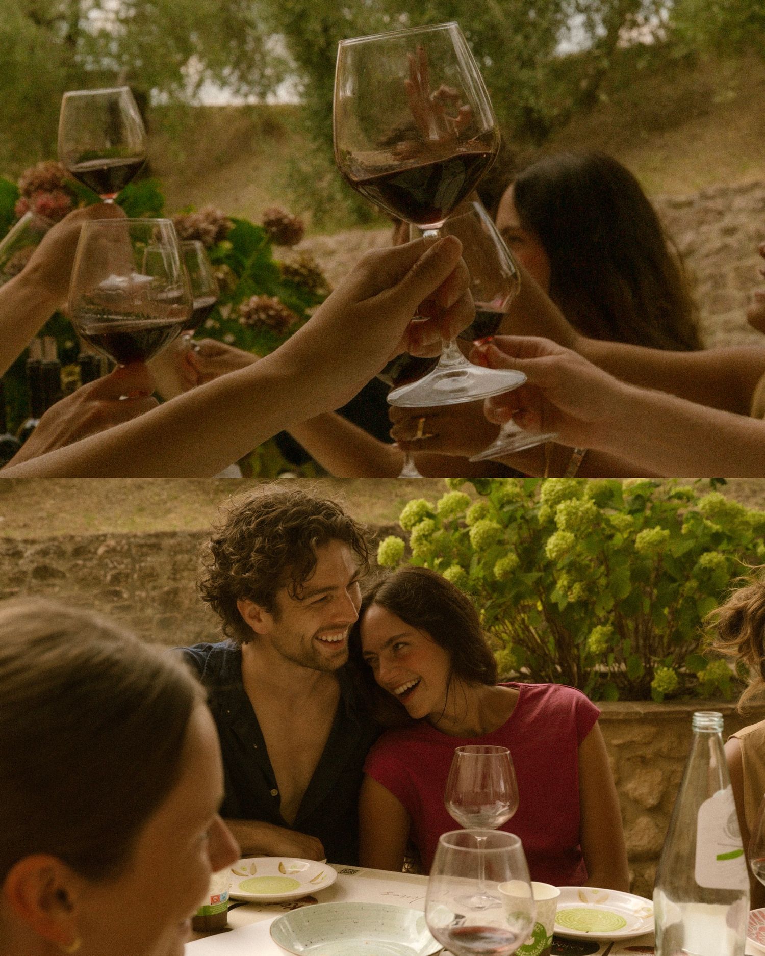 Friends toasting red wine glasses outdoors while a couple smiles intimately at a sunny al fresco dining table.