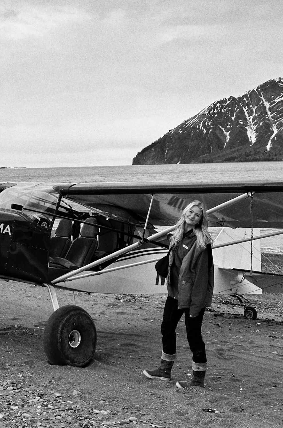 Black and white photo of a woman standing beside a small bush plane on a rocky shore with mountains in the background.