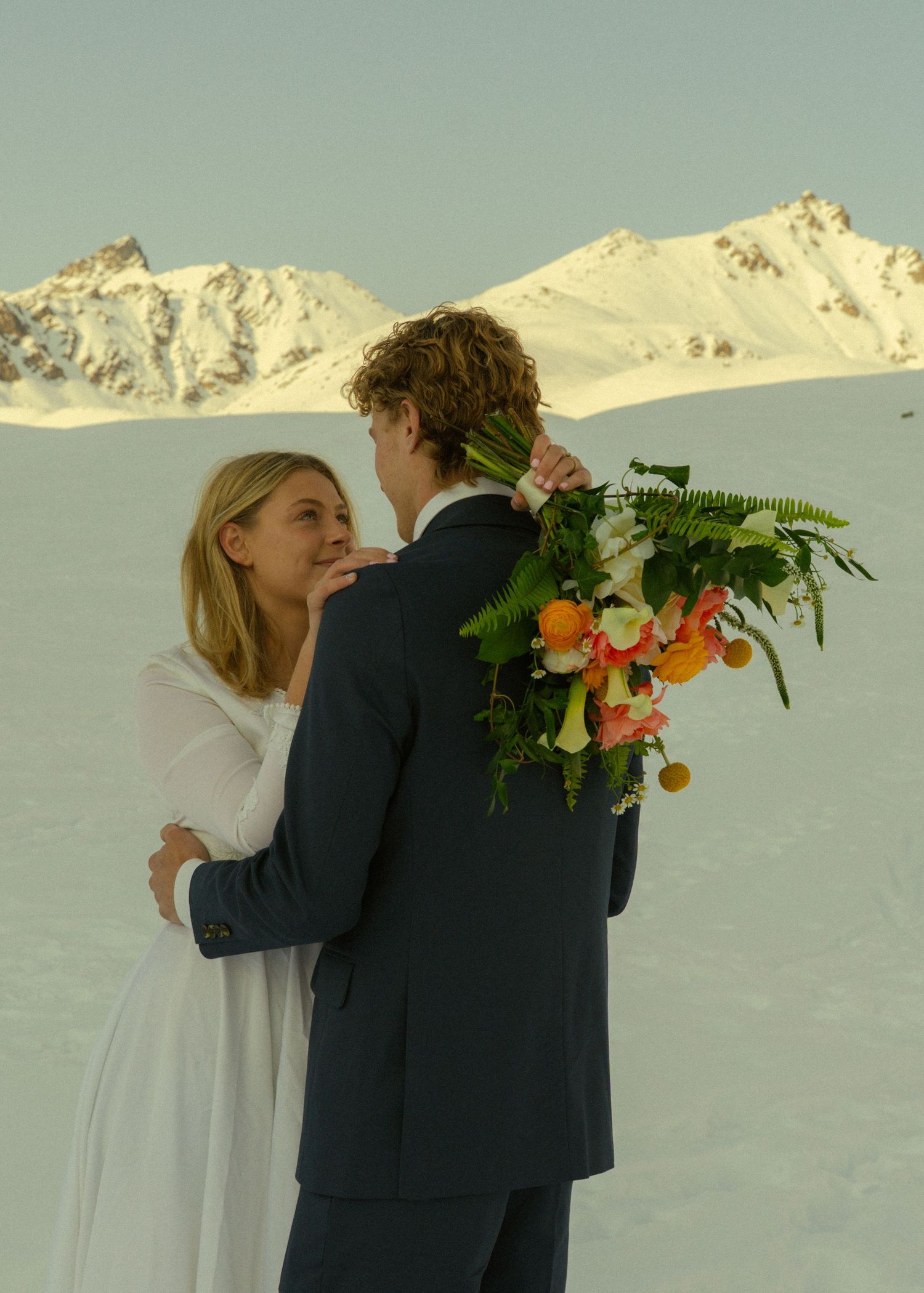 Bride gazing at groom holding vibrant tropical bouquet with stunning snowy mountain range behind them.