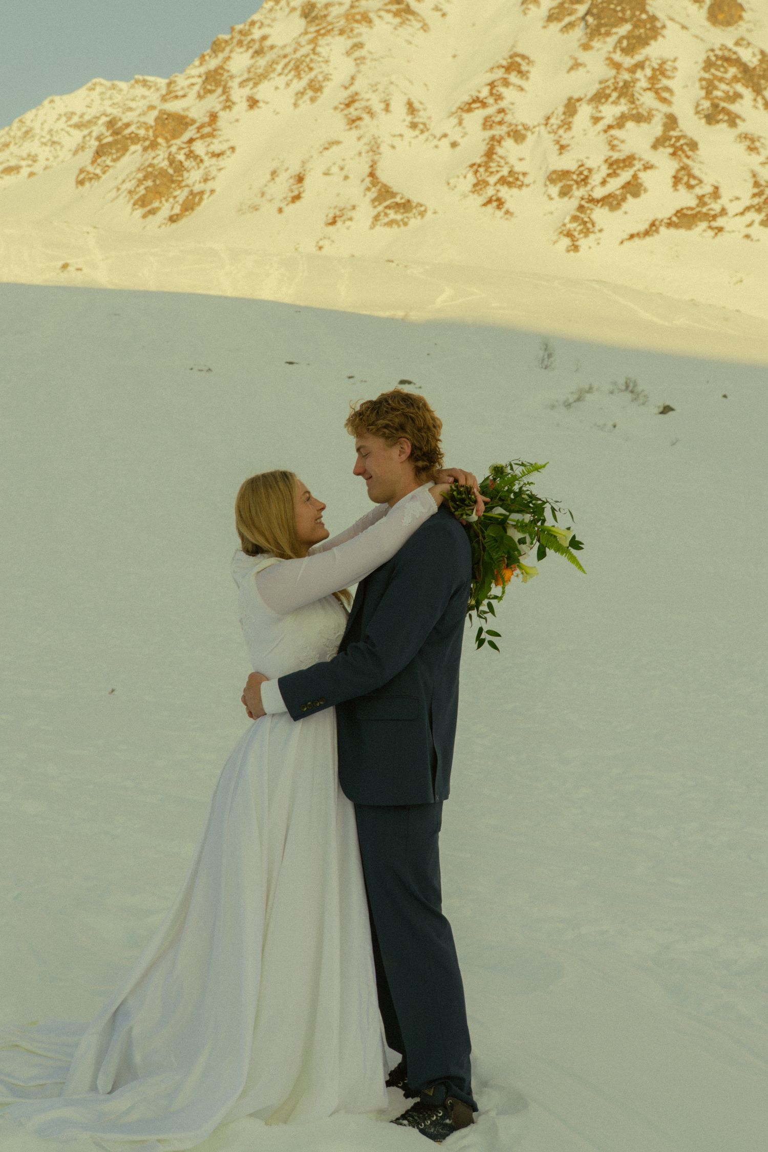 Wedding couple embracing on snow-covered mountain, bride in white gown holding bouquet facing groom in suit.