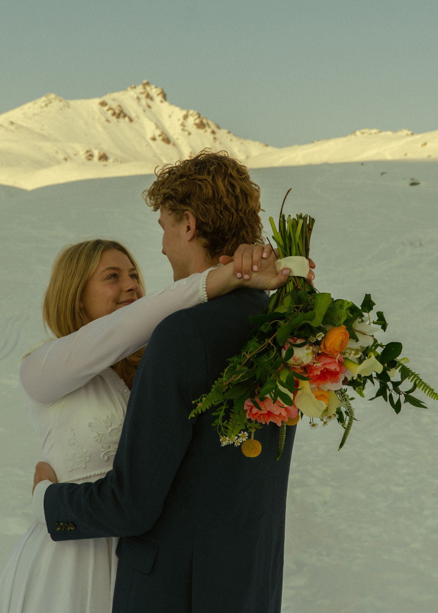 Wedding couple embracing on snowy mountain slope, groom holding lush tropical bridal bouquet at sunset.