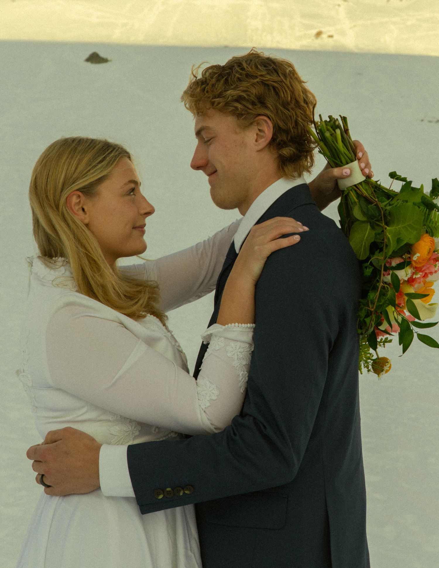 Bride and groom embracing face-to-face in the snow, she in white lace dress, he in navy suit holding orange bouquet.