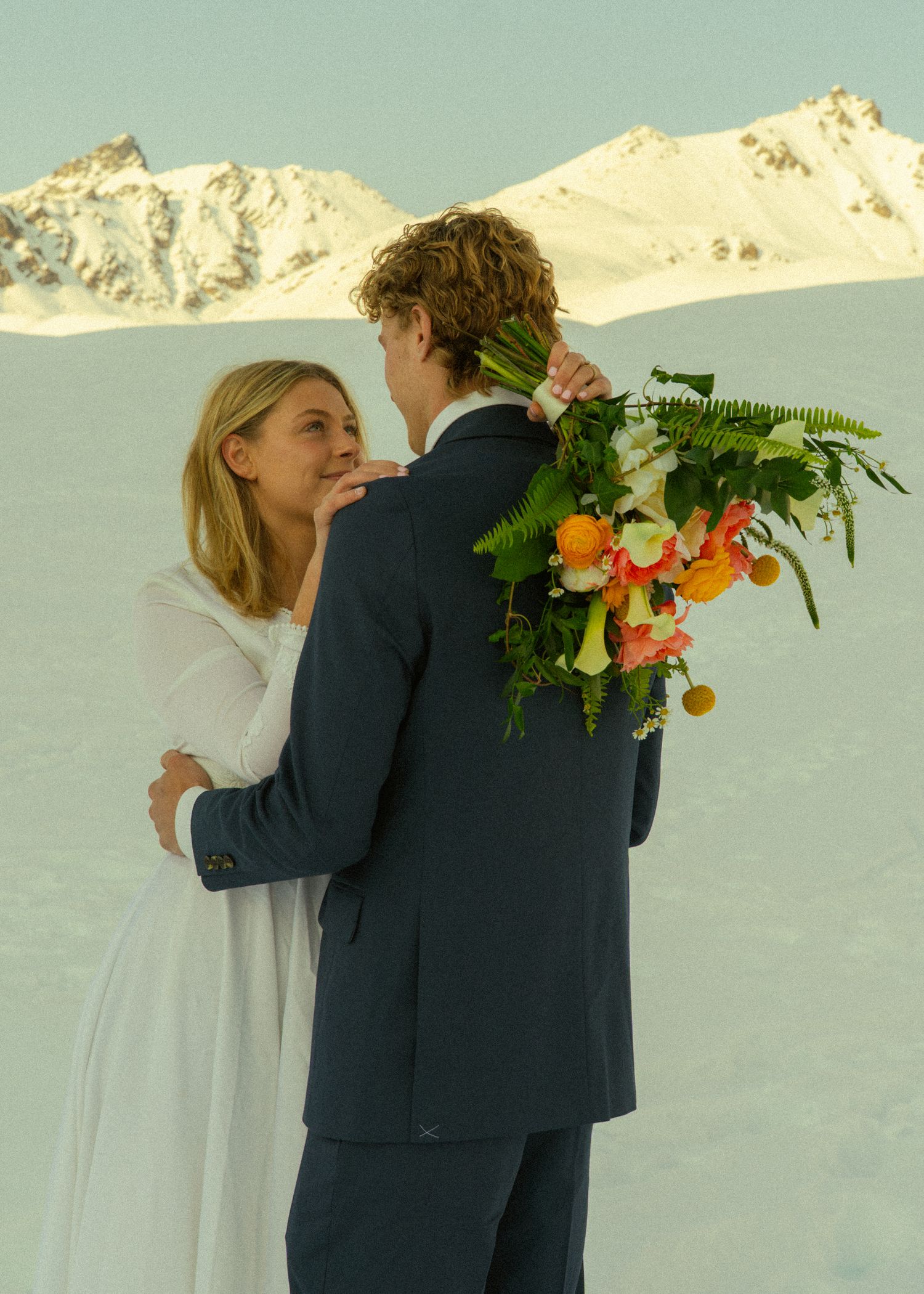 Smiling bride in white dress embraces groom holding tropical bouquet against snow-covered mountain backdrop.