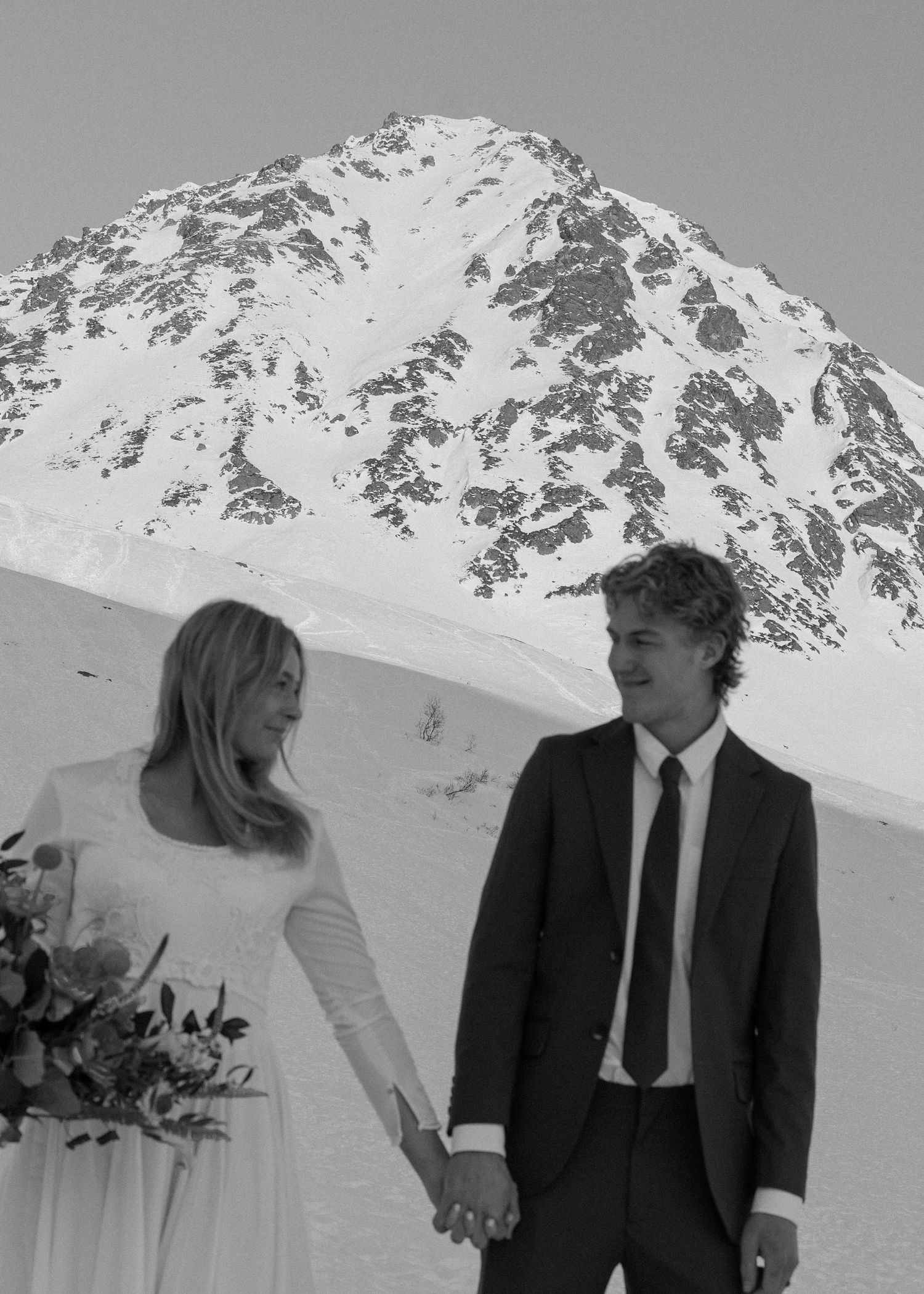 Black and white photo of a bride and groom holding hands with a snowy mountain peak behind them.