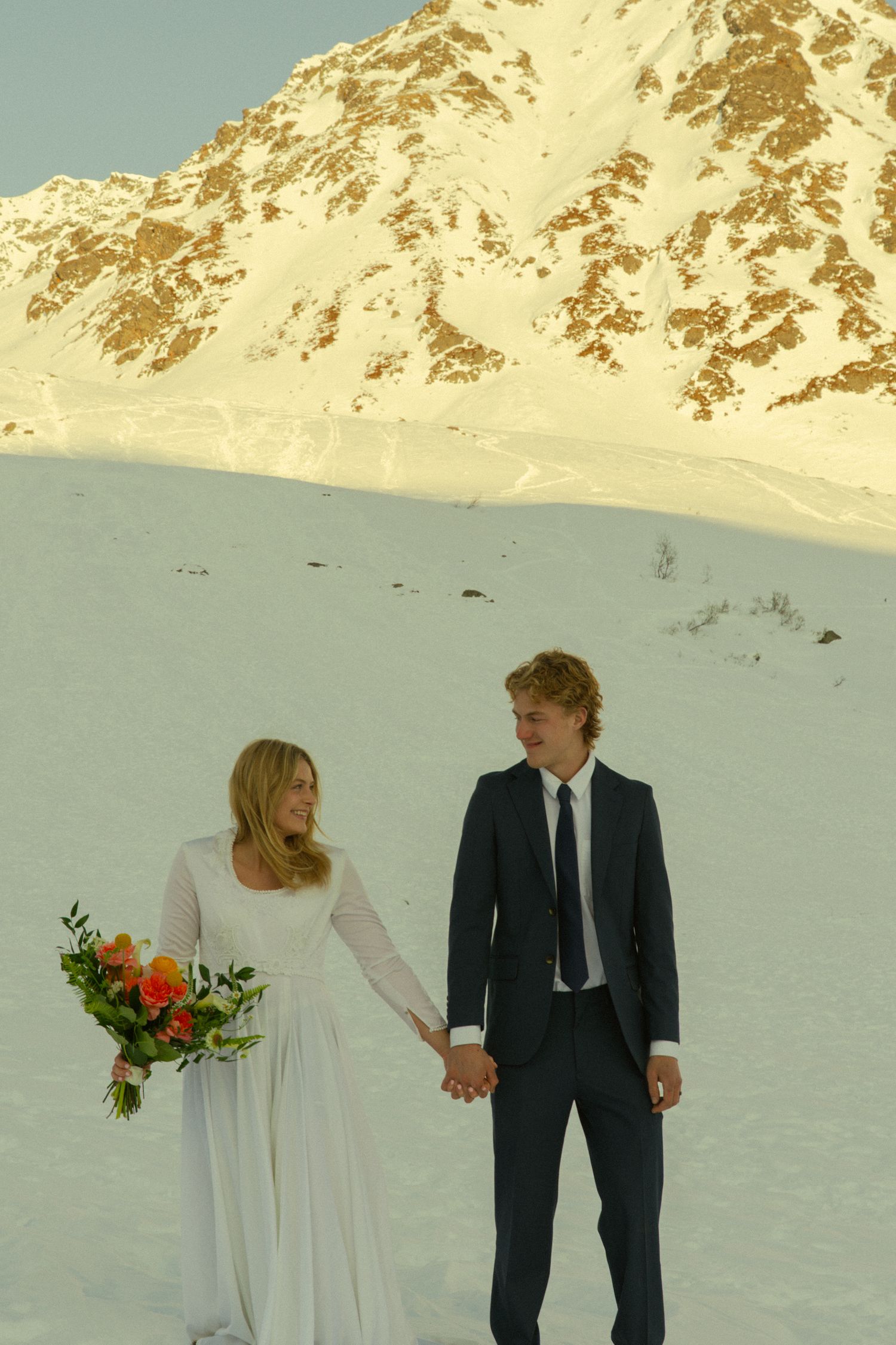 Bride and groom holding hands in white dress and navy suit with colorful bouquet against a snowy mountain landscape.