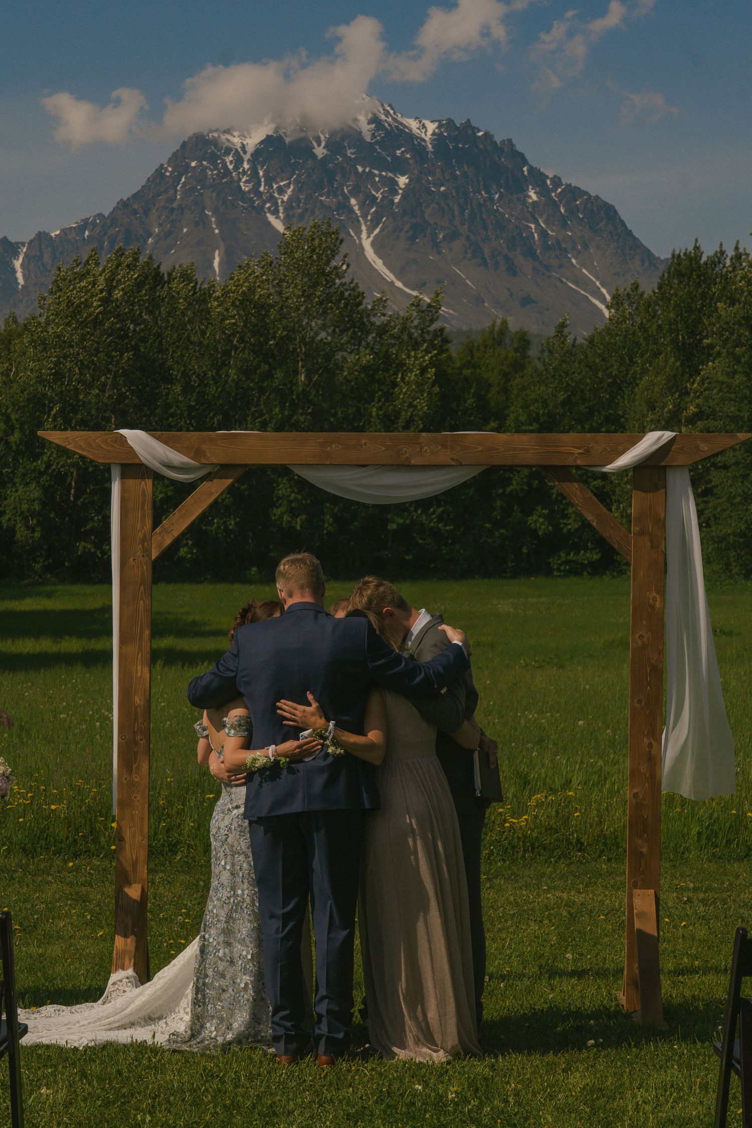 Wedding party shares group hug under rustic wooden arch with dramatic snow-capped mountain in background.
