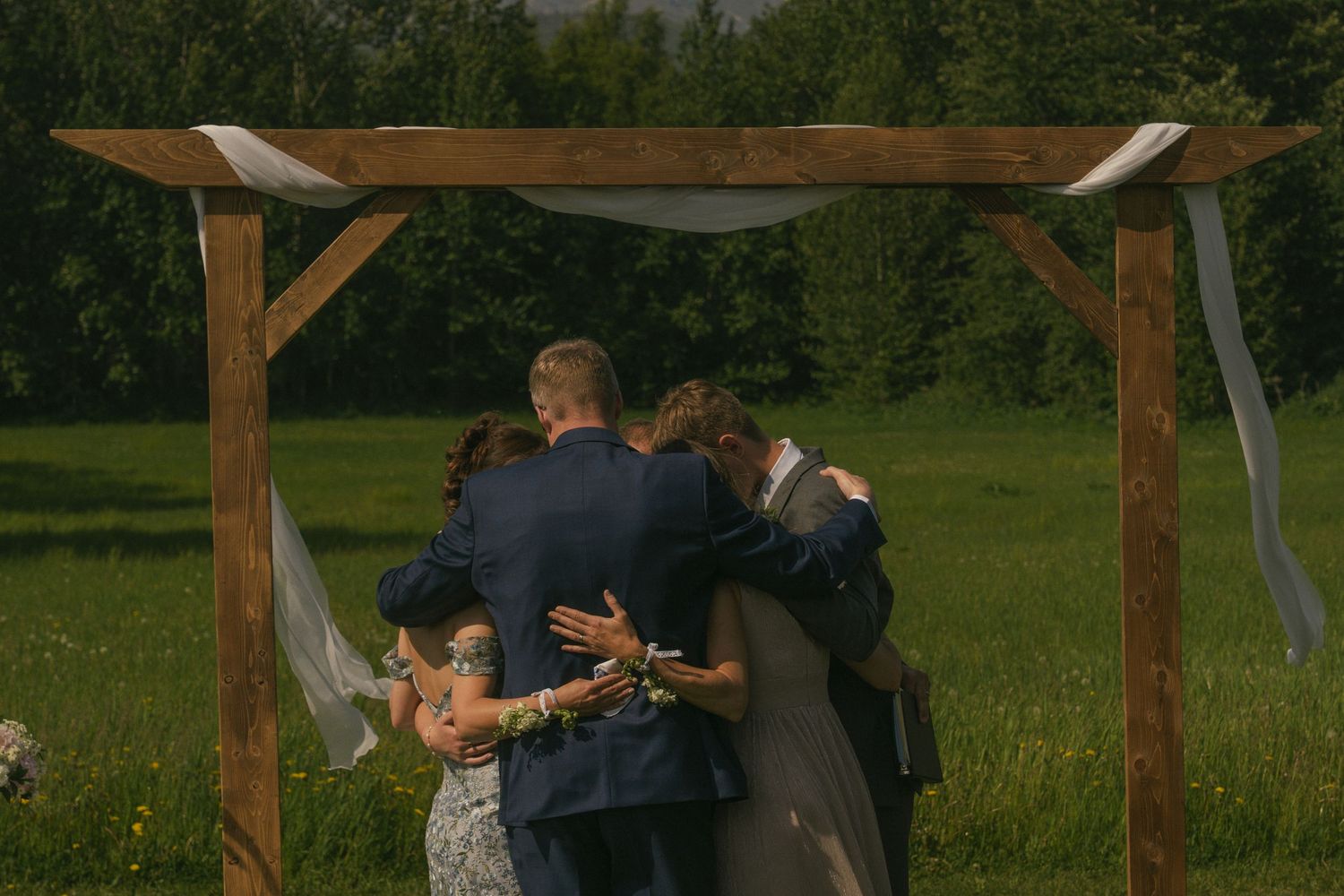 Wedding party shares intimate group hug under rustic wooden arch in lush green outdoor setting.