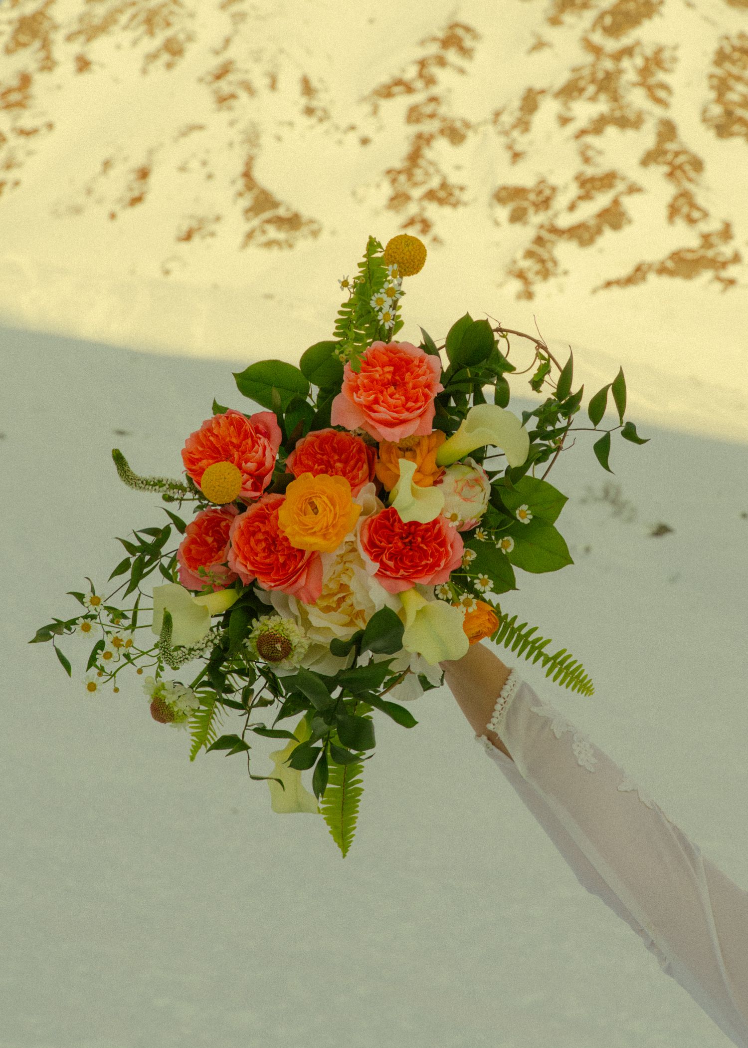Lush bridal bouquet of orange roses and greenery held by bride in white dress on snowy terrain.
