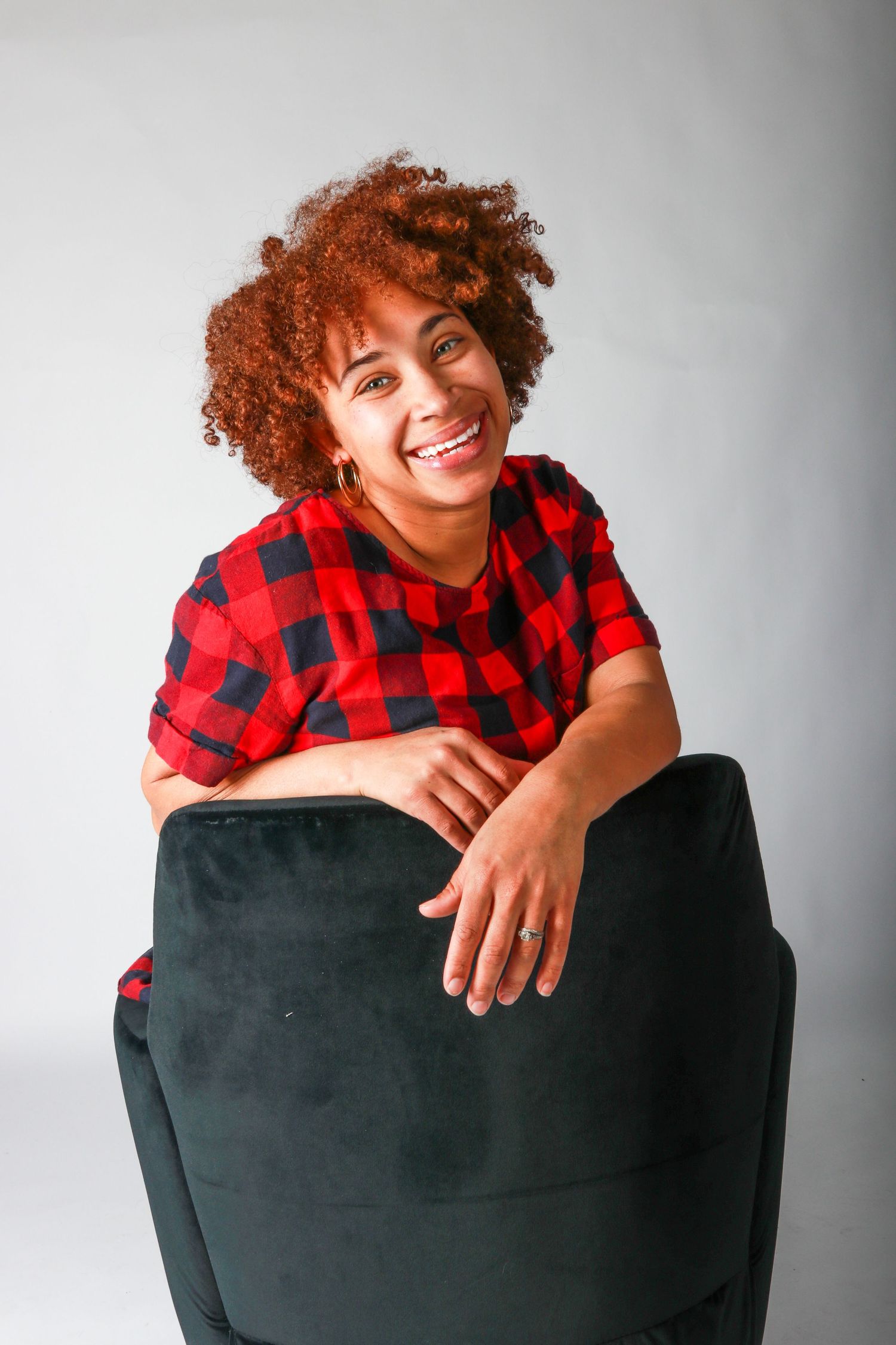Smiling woman with curly red hair in a red plaid shirt leaning on a black velvet chair.