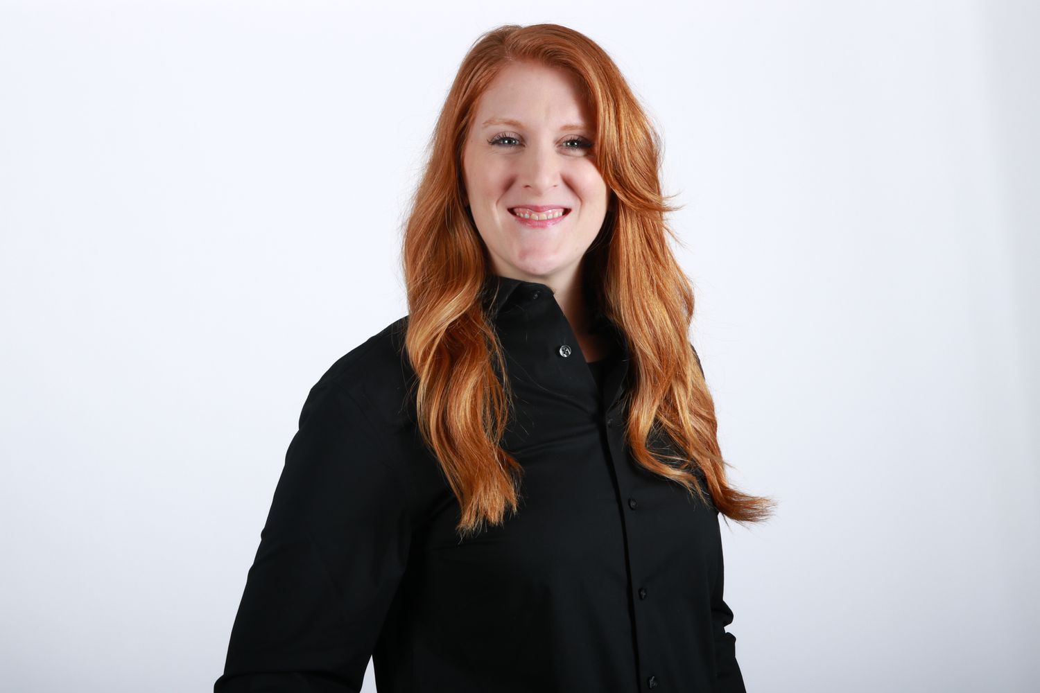 Smiling woman with long red hair wearing black shirt poses for professional headshot on white background.