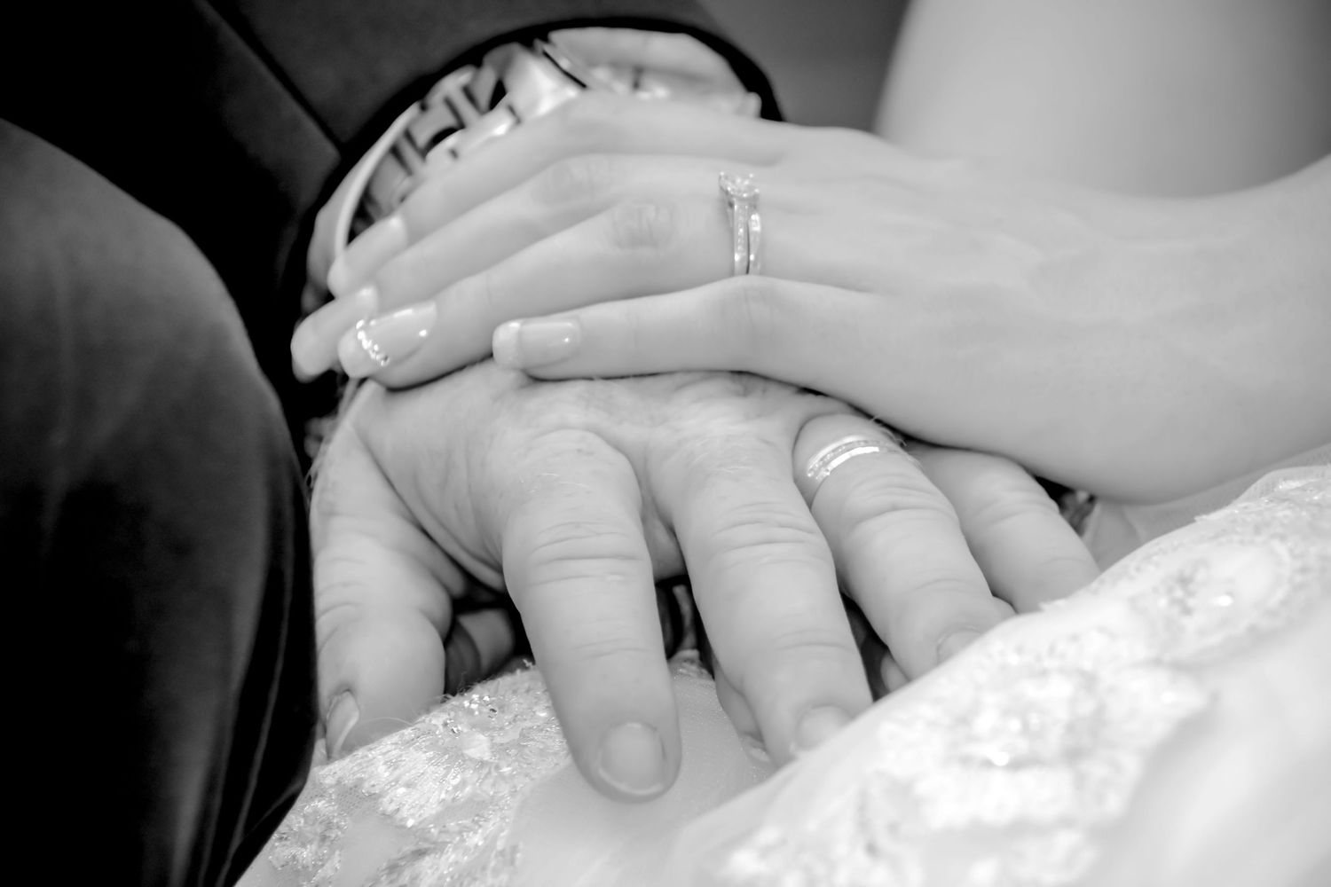 Black and white close-up of newlyweds hands stacked together showing wedding rings on lace dress.
