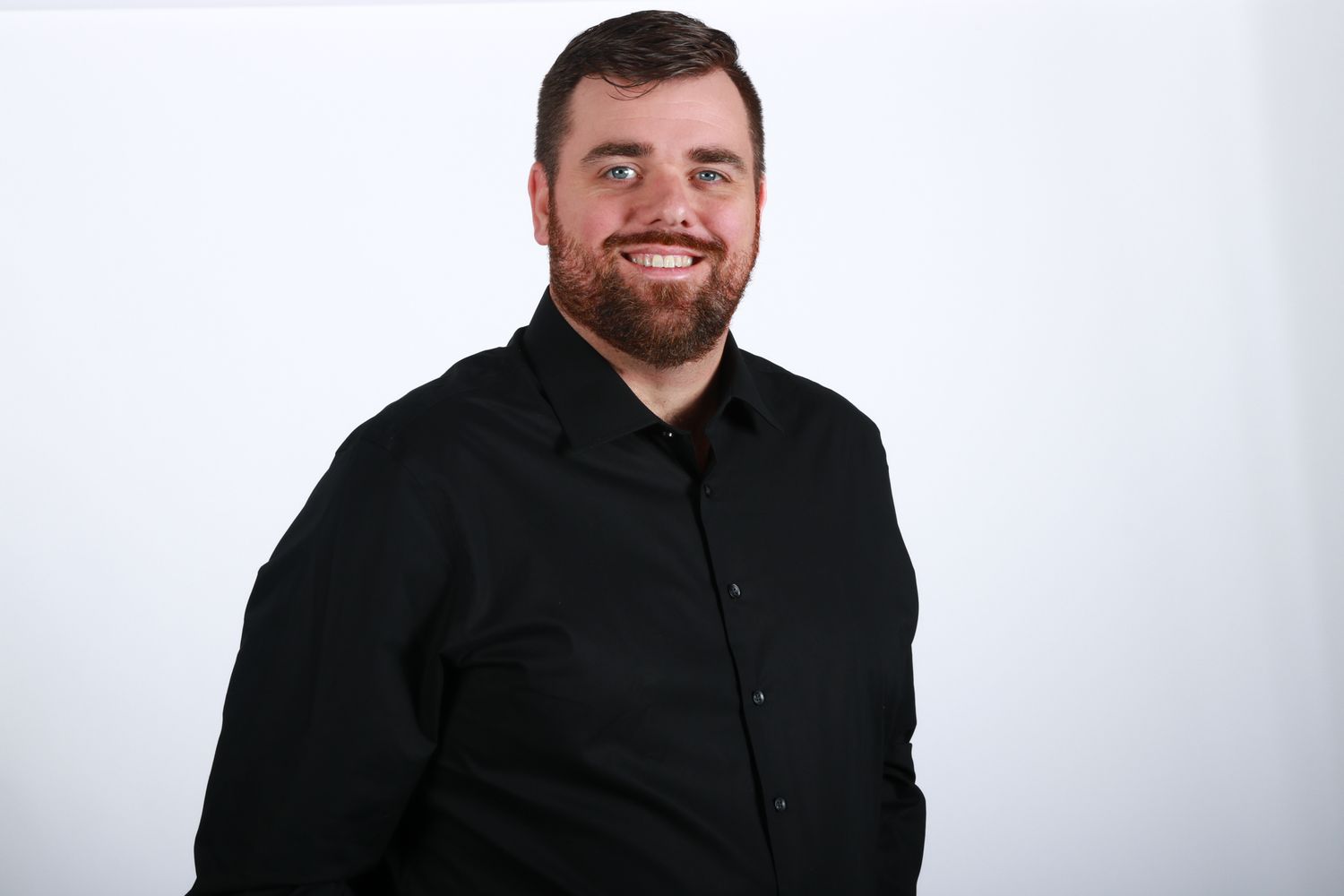 Young bearded man in black dress shirt smiles confidently for professional headshot on white background.