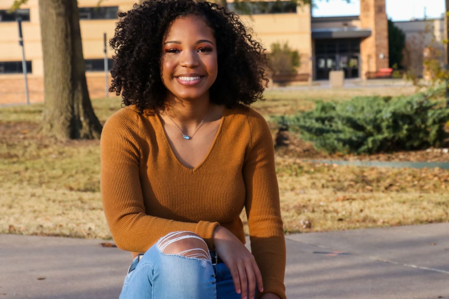 Young Black woman with curly hair smiling outdoors, wearing a mustard sweater and ripped jeans on a sunny day.
