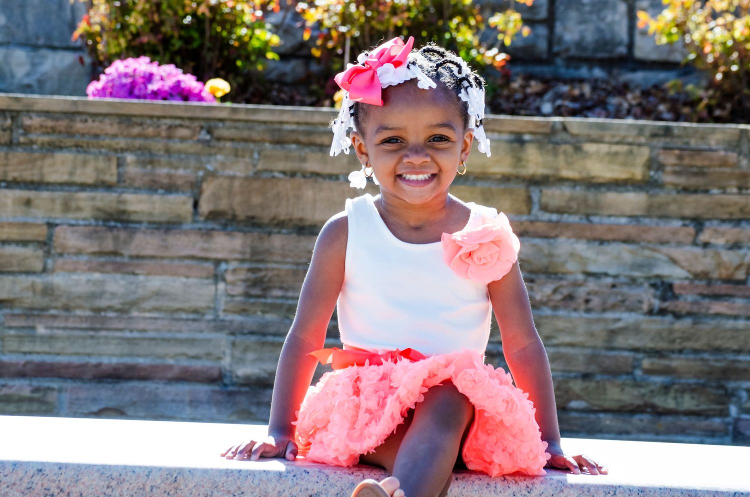 Happy young Black girl in coral tutu skirt and white top sitting outdoors smiling.