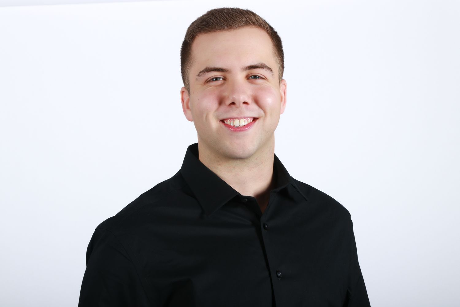 Young smiling man in black button-down shirt poses for clean professional headshot on white background.