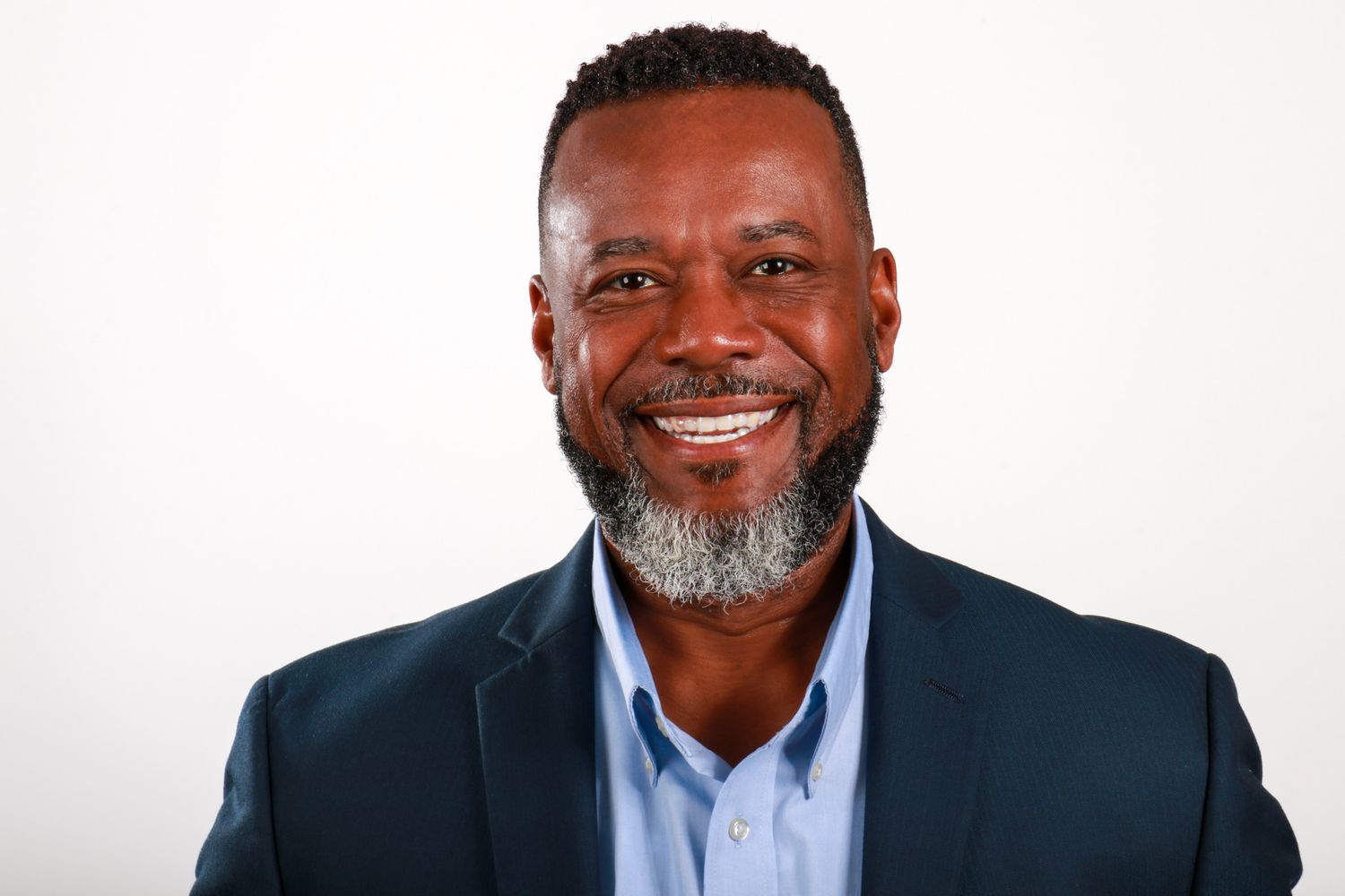 Confident middle-aged Black man with gray beard smiles in navy blazer for professional headshot portrait.