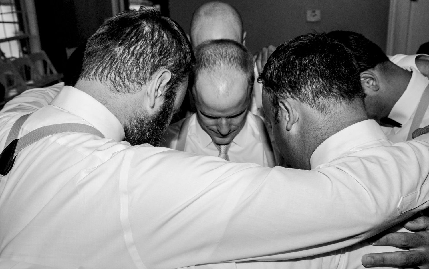 Black and white photo of groomsmen in white shirts huddled together in a prayer circle at a wedding.