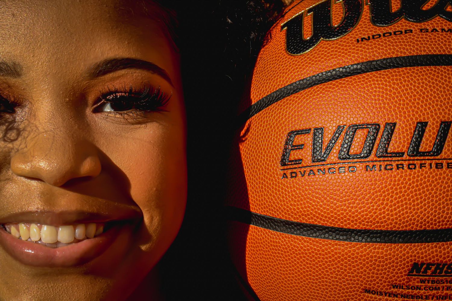 Close-up portrait of a smiling young woman beside a Wilson Evolution basketball in dramatic lighting.