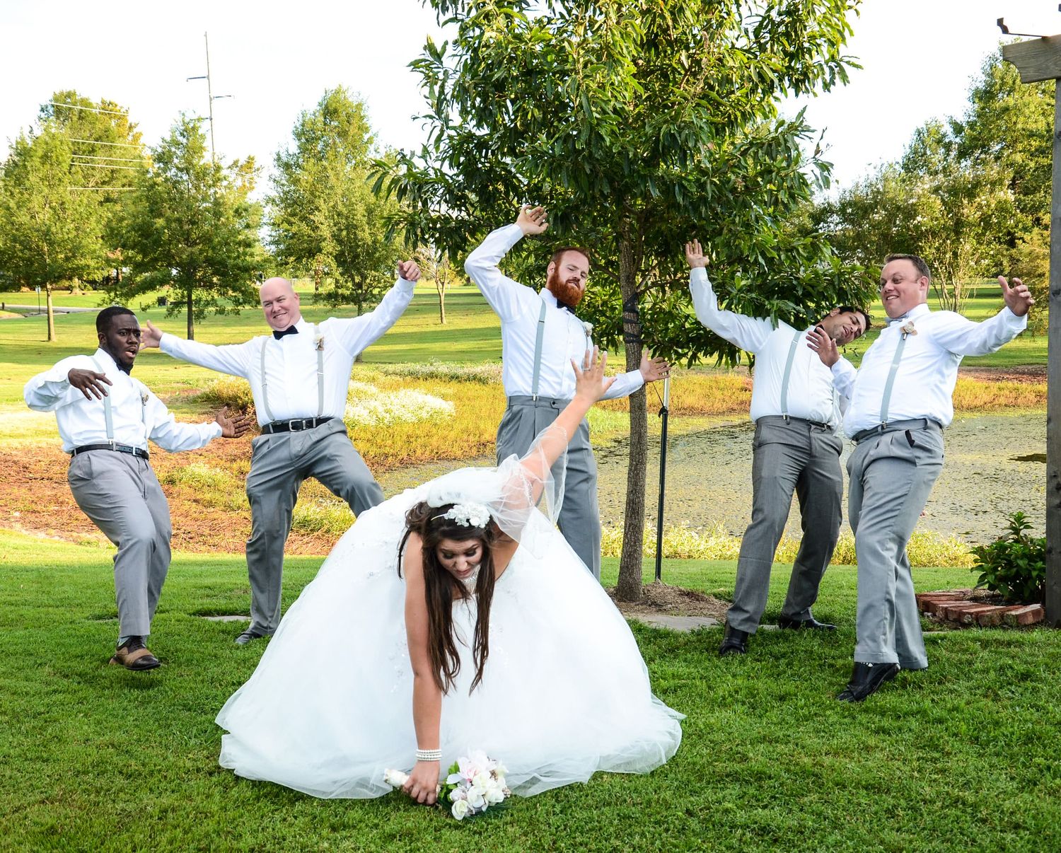 Bride in white ball gown kneels while groomsmen in gray suits pose playfully around her outdoors.