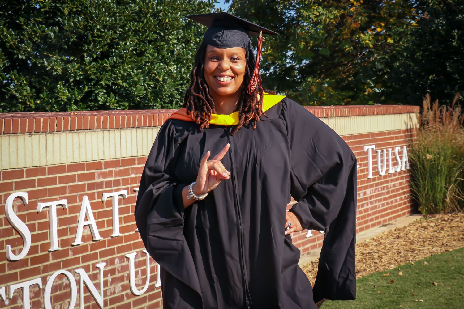 Woman in graduation cap and gown posing in front of Tulsa State sign outdoors.