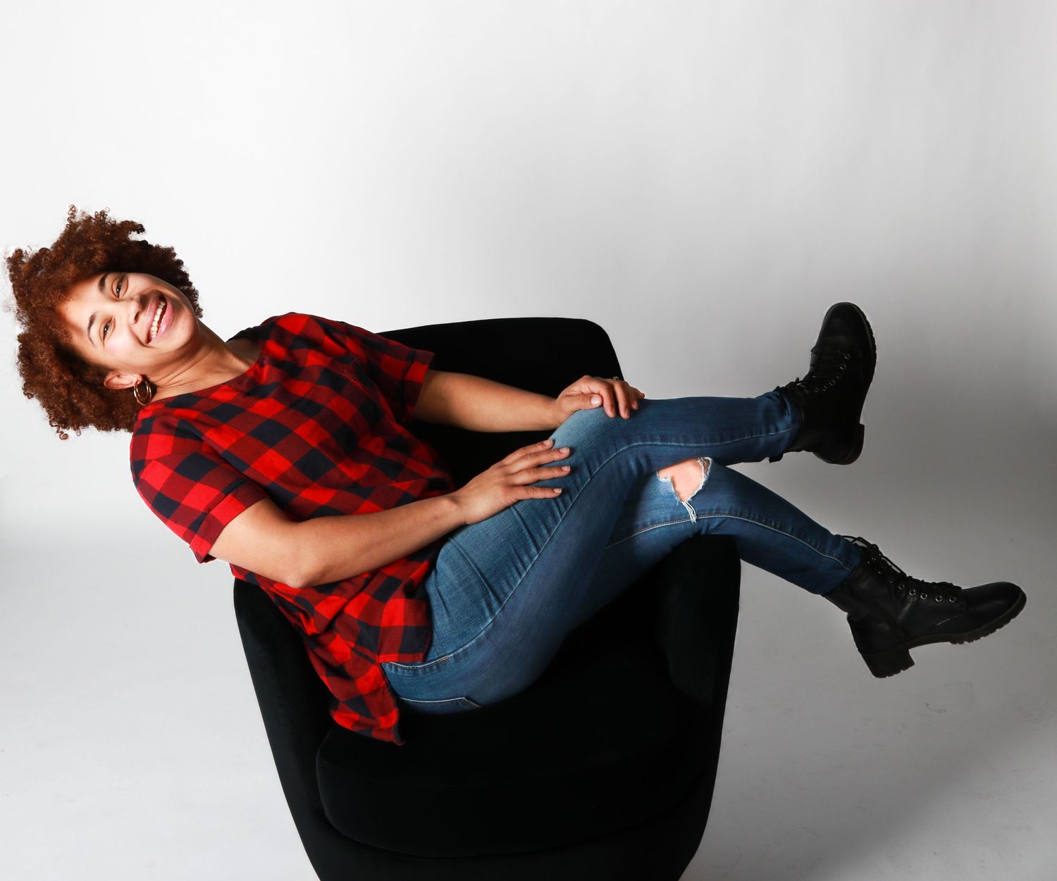 Young woman in red plaid shirt and jeans lounging in a black chair laughing.