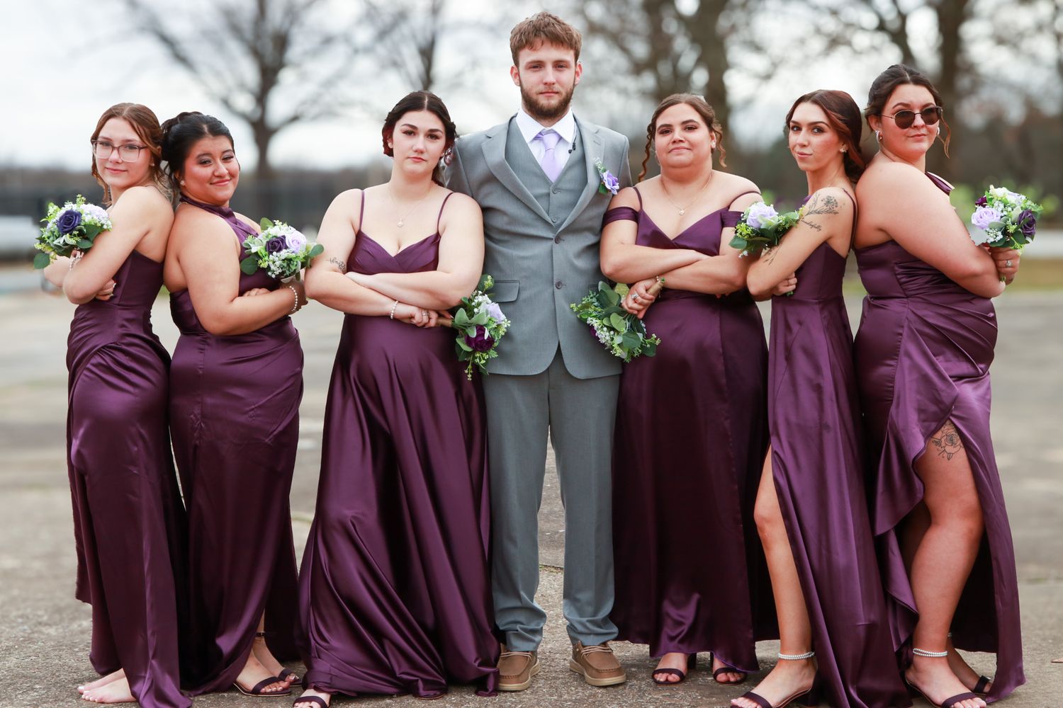 Groomsman in gray suit poses with six bridesmaids in purple satin gowns holding bouquets at outdoor wedding.