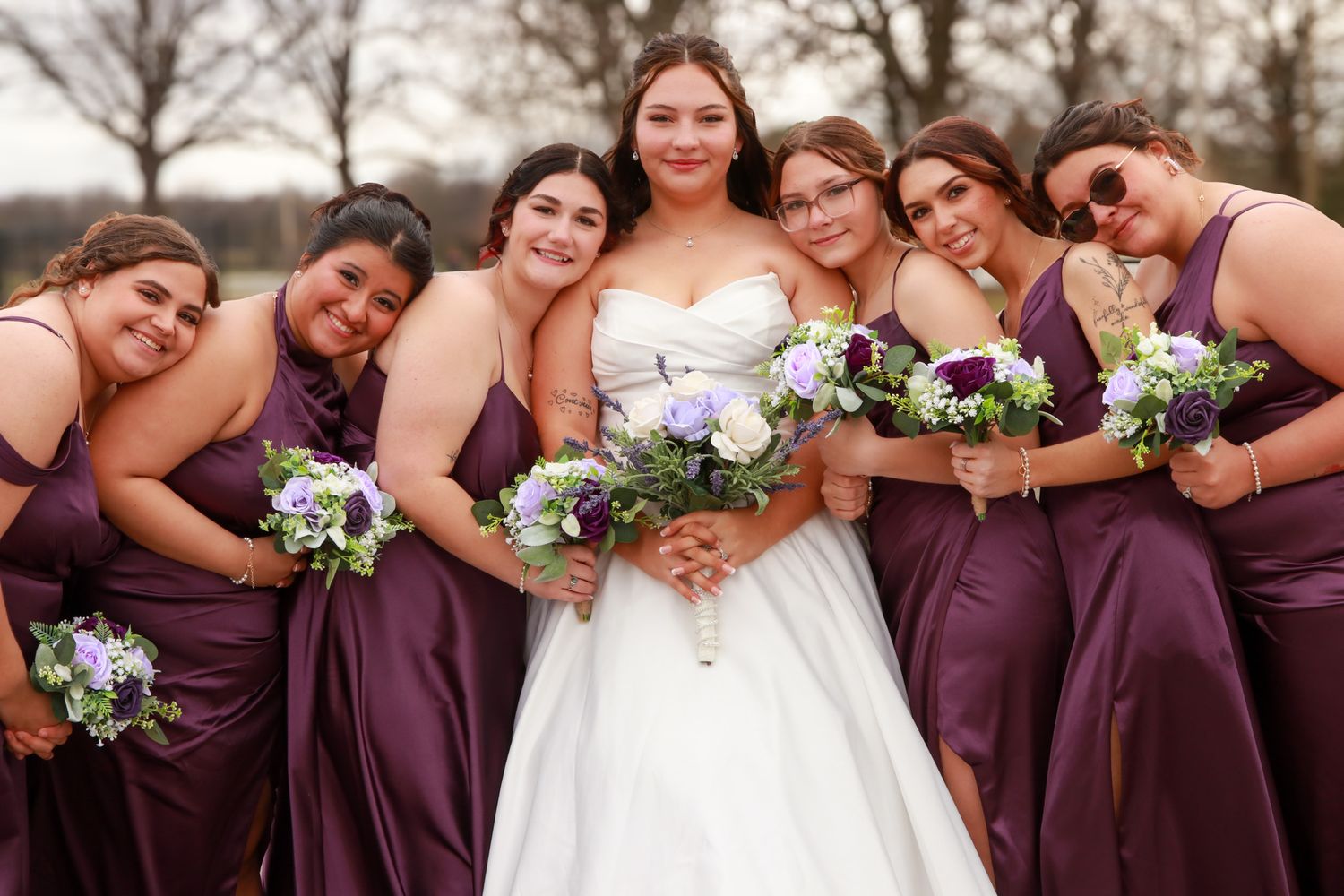 Bride surrounded by six smiling bridesmaids in purple dresses holding bouquets.
