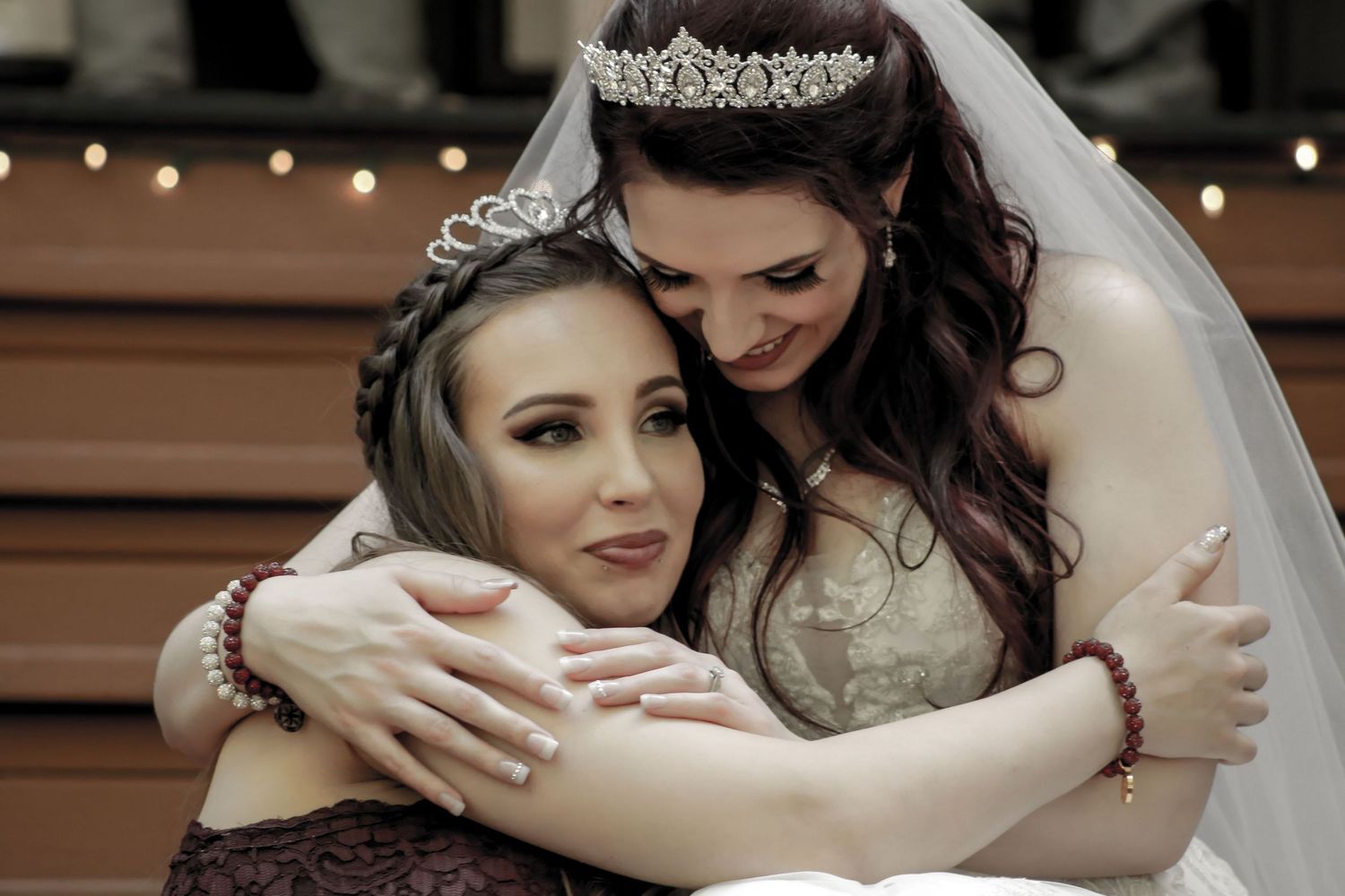 Bride in white gown and tiara shares tender hug with bridesmaid in purple dress at wedding celebration.