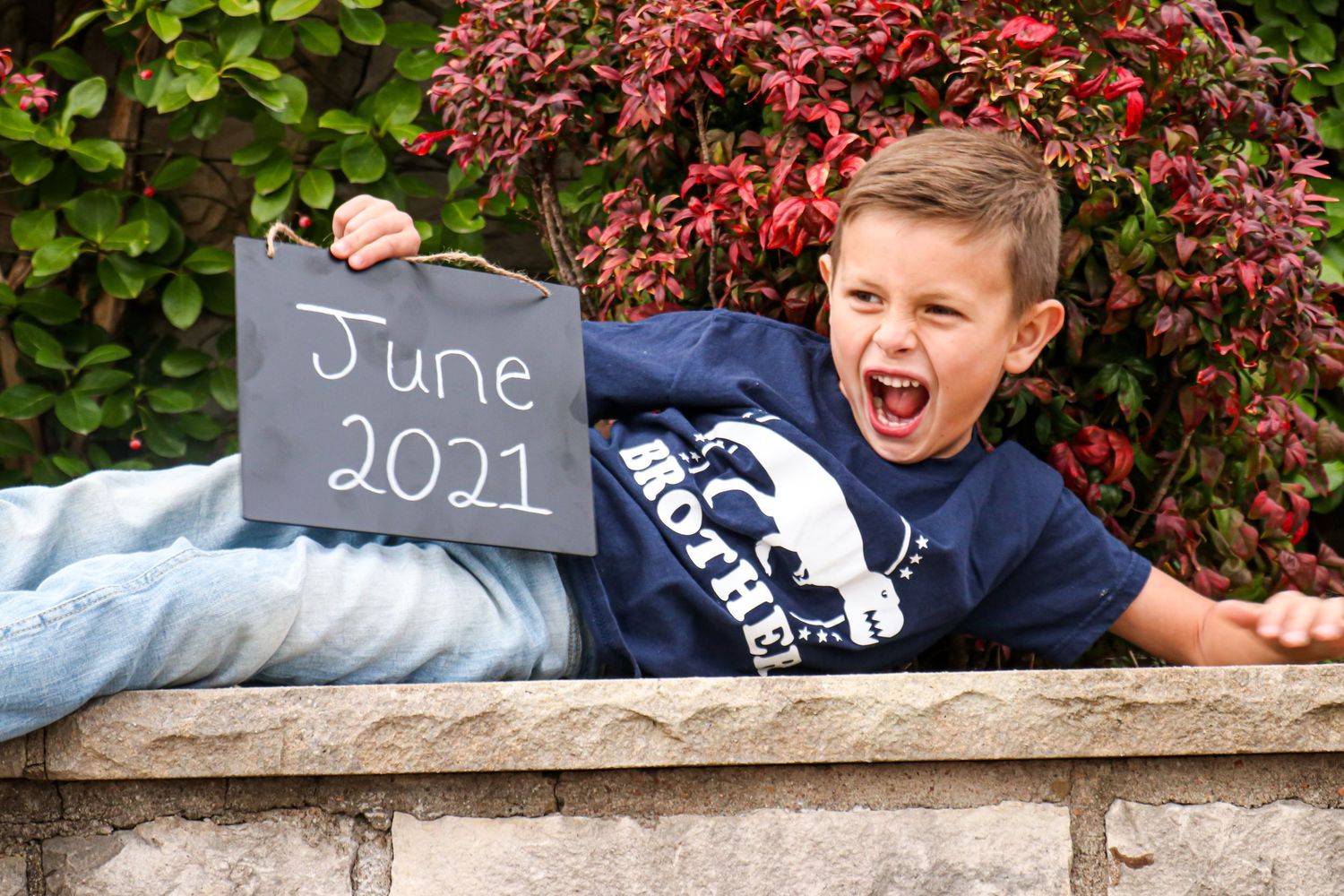 Excited young boy holding a June 2021 chalkboard sign while lying on a stone wall.
