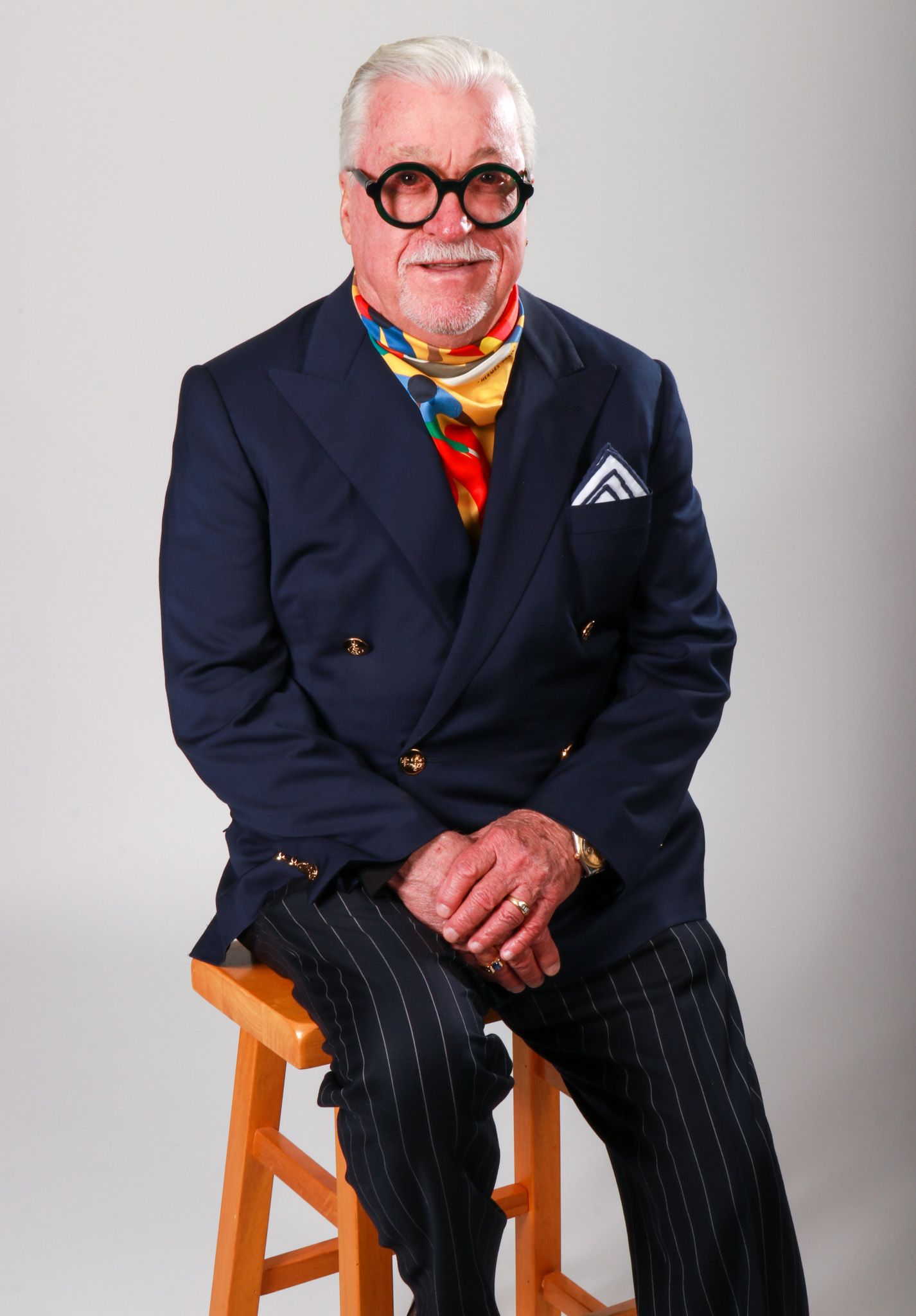 Stylish elderly man in navy pinstripe suit with colorful scarf and bold glasses sits on stool.