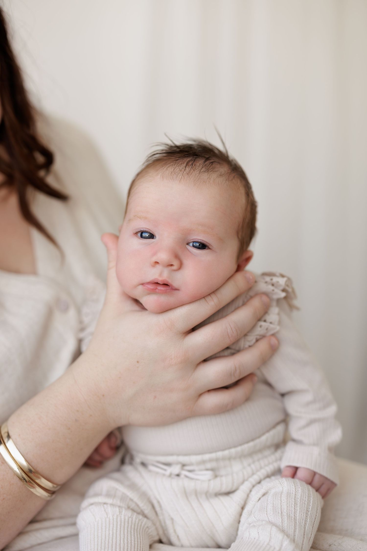 Baby in cream-colored outfit supported by gentle hands against white backdrop.