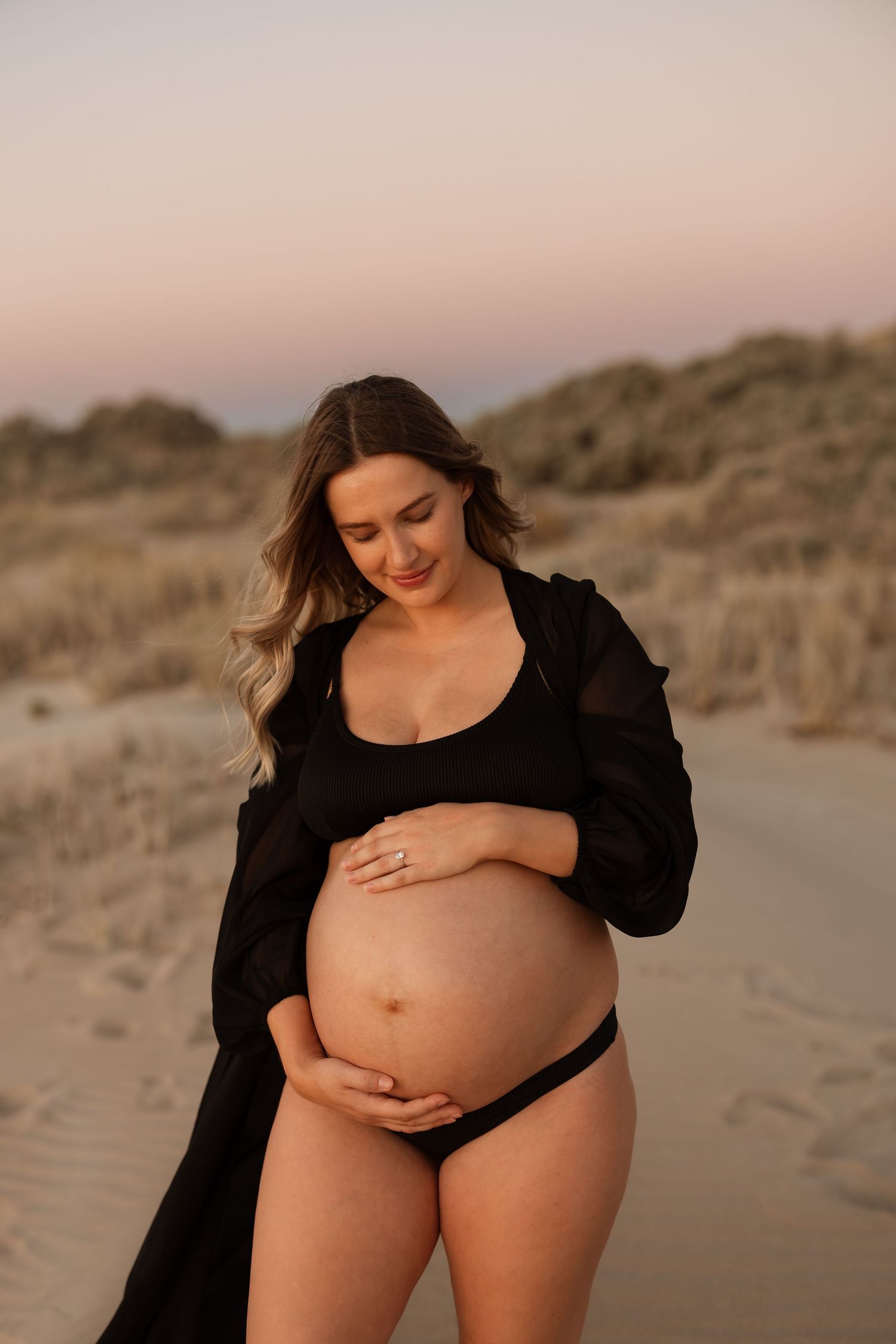 Artistic maternity photo of a pregnant figure in black attire against sand dunes during golden hour.