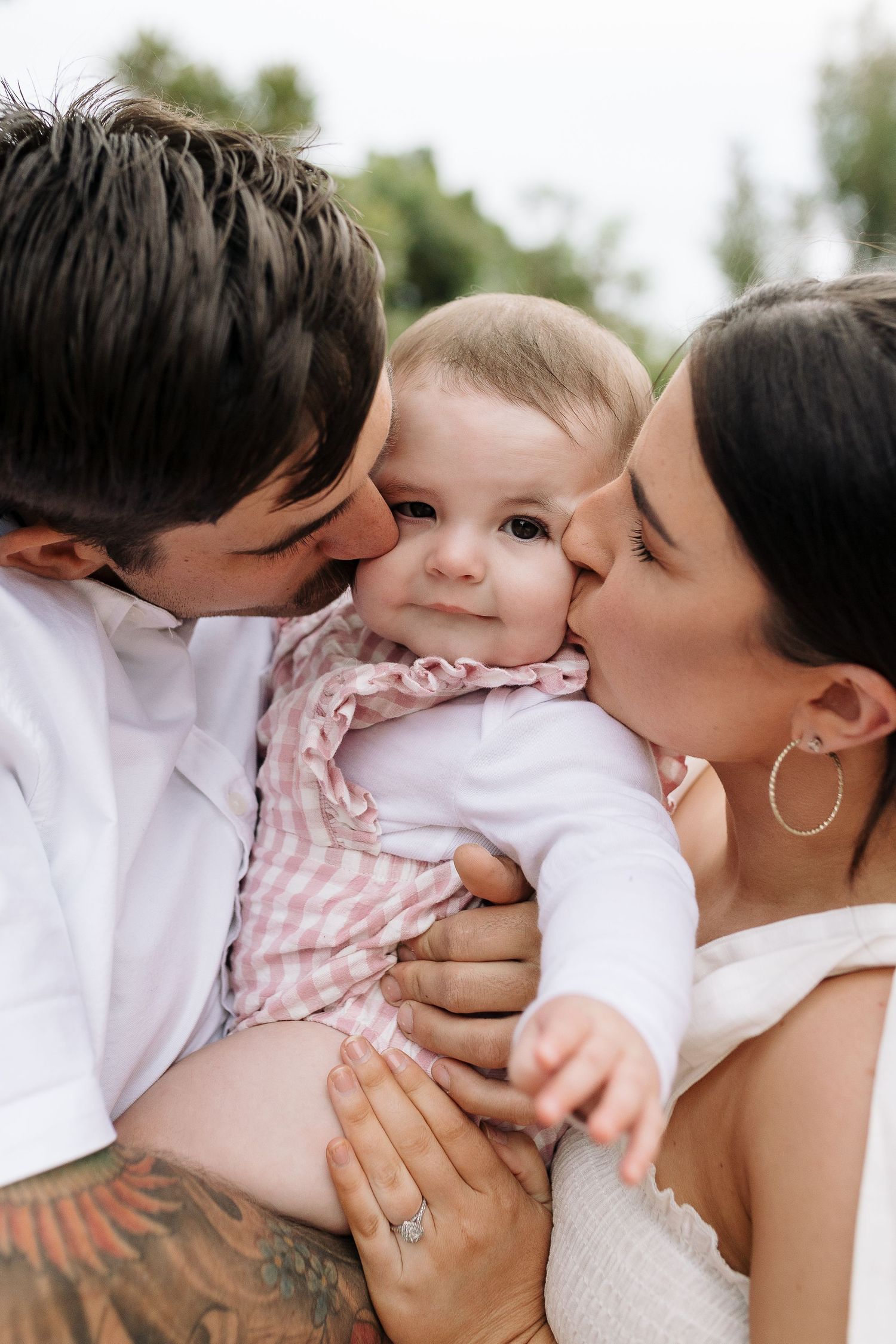 Intimate family moment outdoors showing parents kissing baby wearing pink and white.