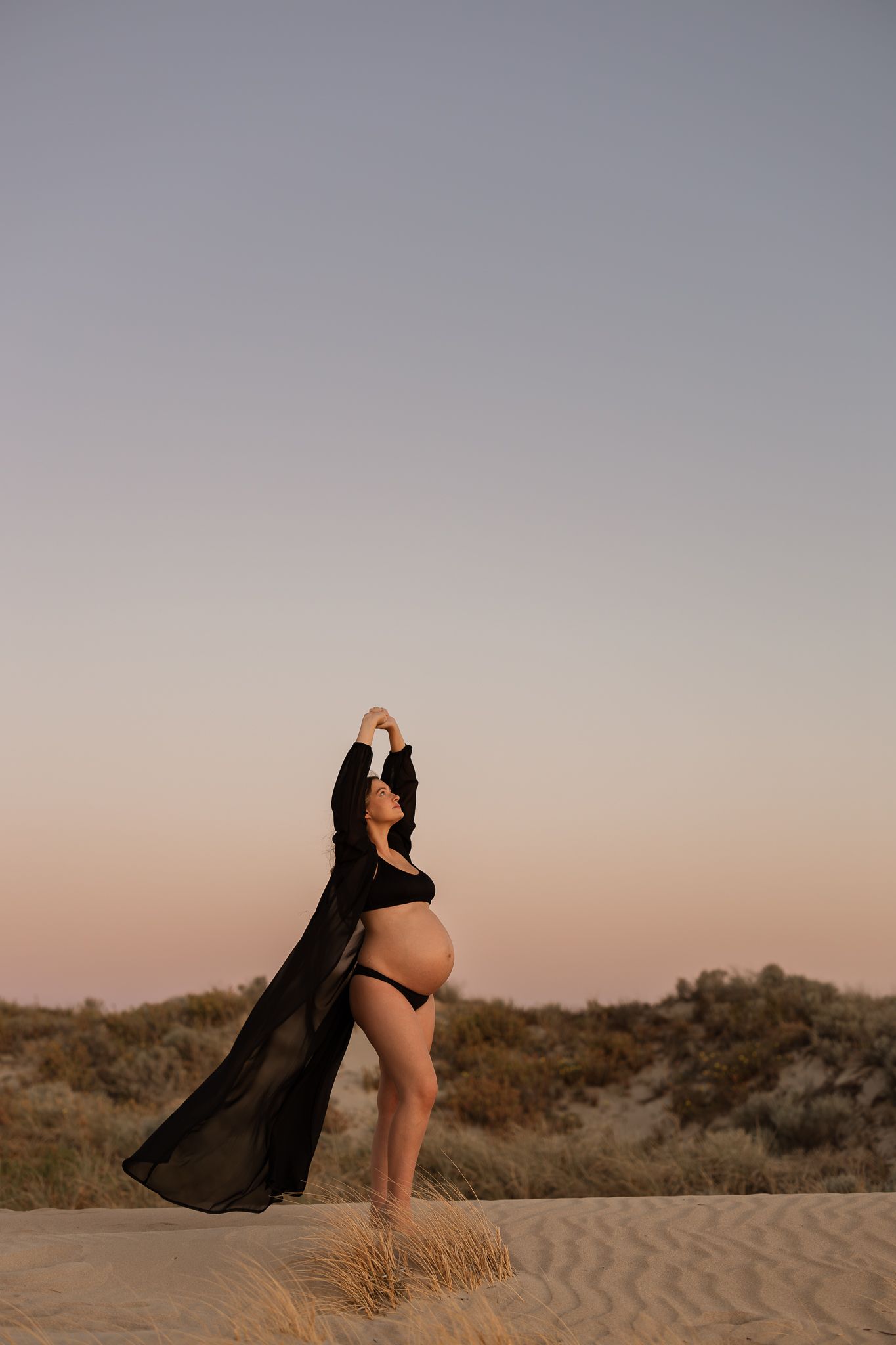 Silhouette of a person in flowing black fabric posing dramatically on sand dunes at sunset.