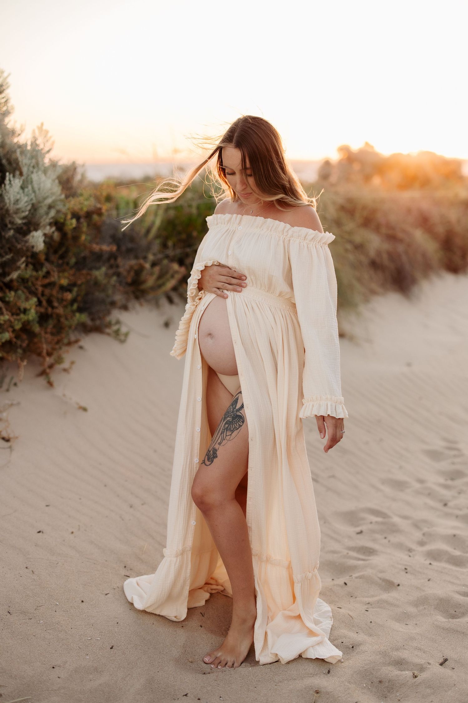 Maternity photo session at sunset on sandy beach with flowing cream-colored dress and natural lighting.