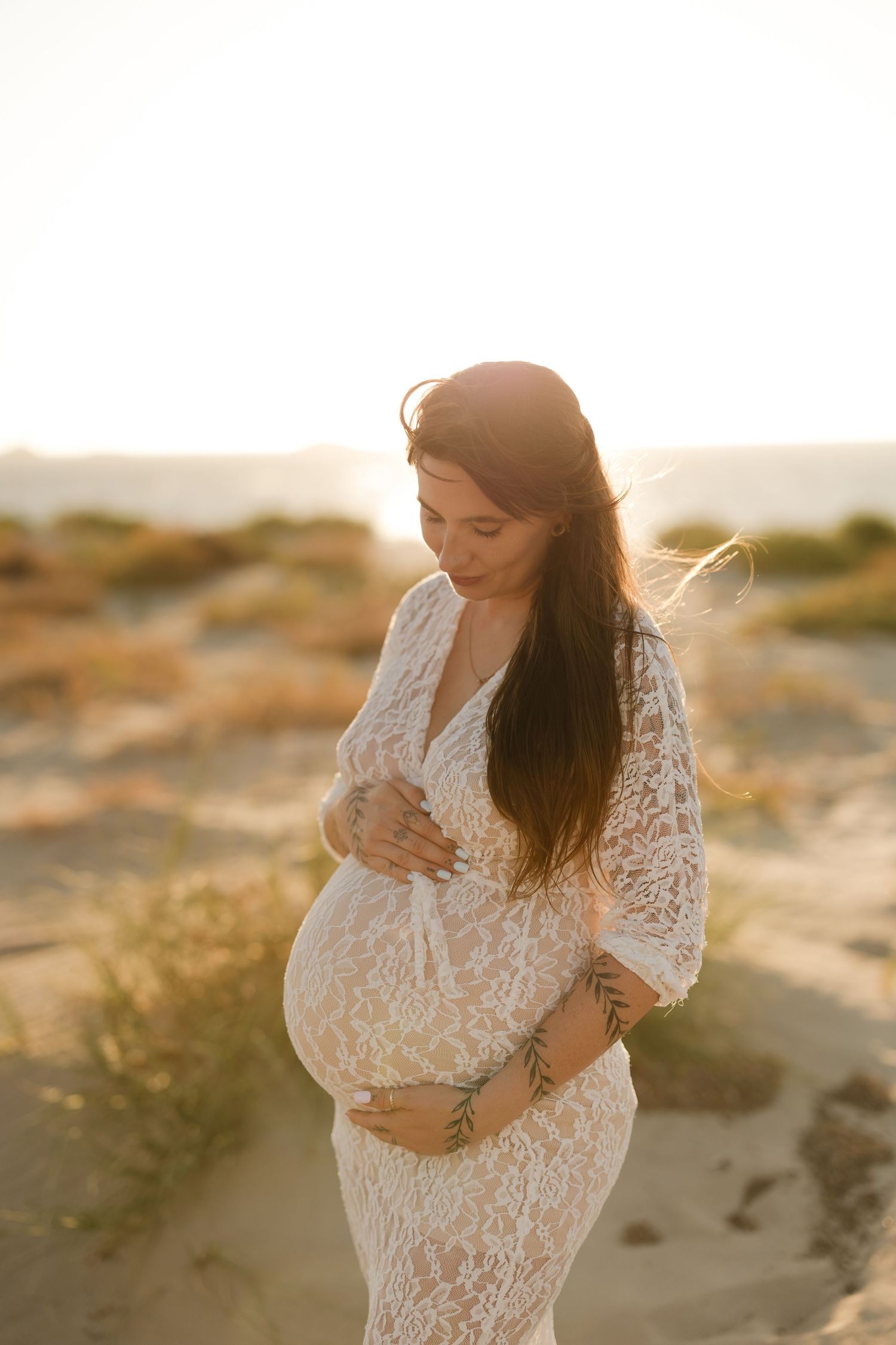 Expectant mother in white lace dress posing on beach at golden hour.