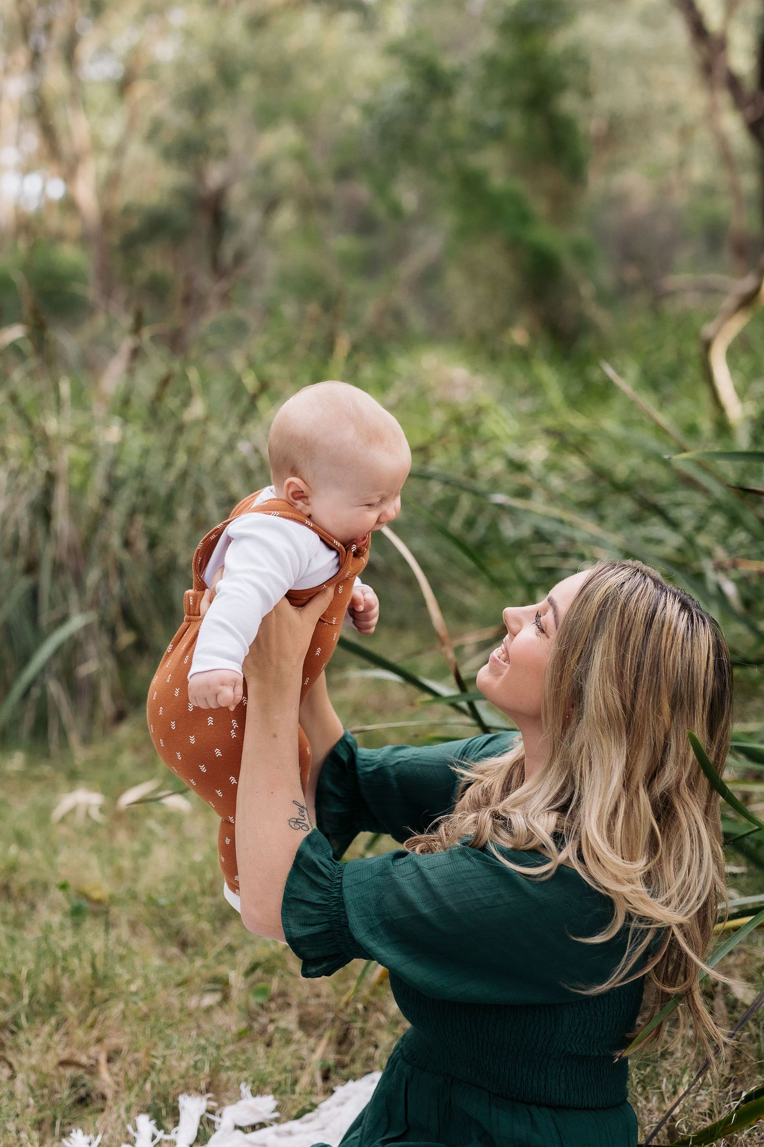 A woman in green sweater lifts baby wearing brown overalls against natural outdoor backdrop.