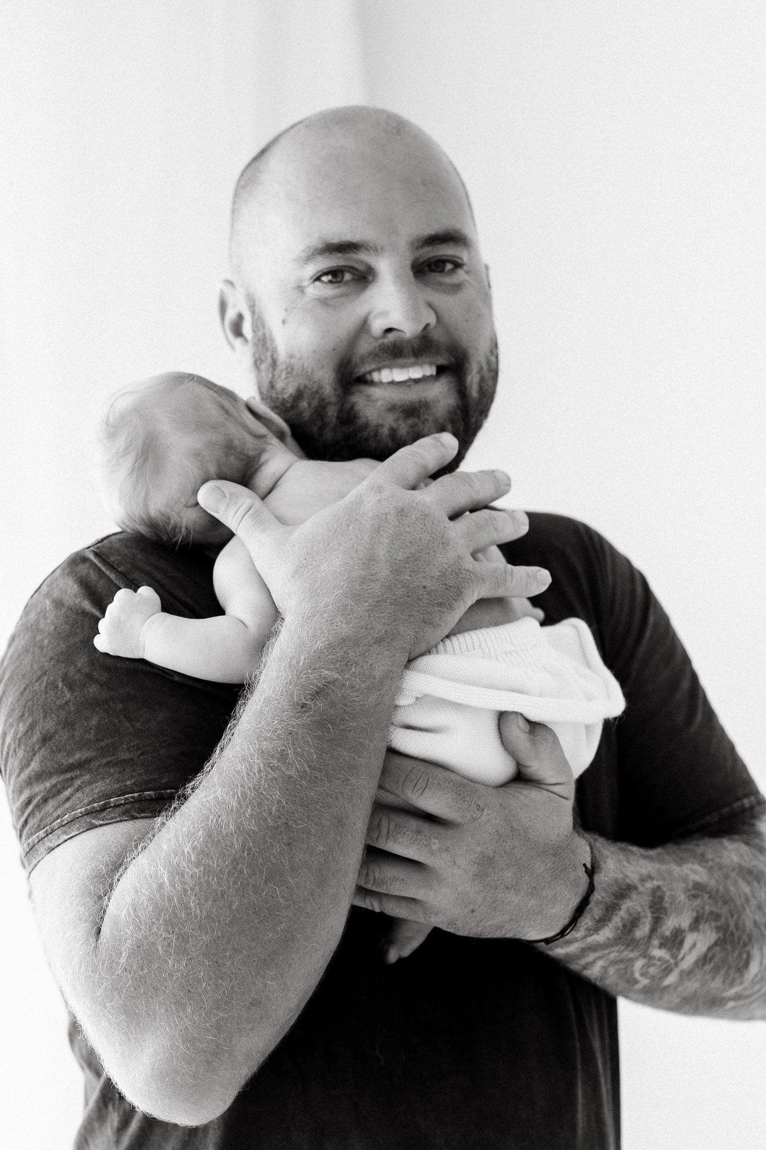 Black and white portrait of a bearded man in dark shirt cradling a newborn while smiling warmly.
