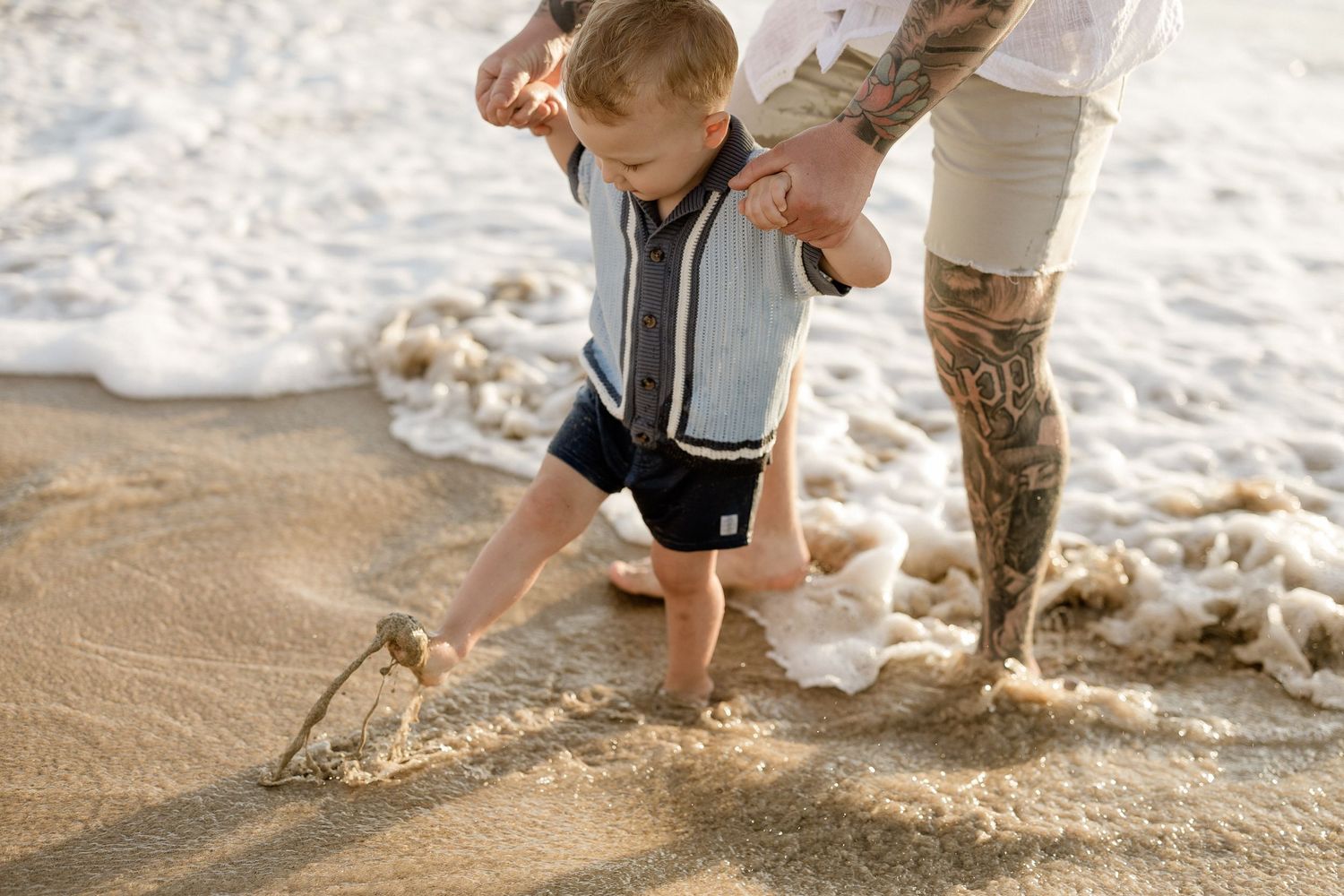 A small child plays in the ocean waves while holding an adult's hand on the beach.