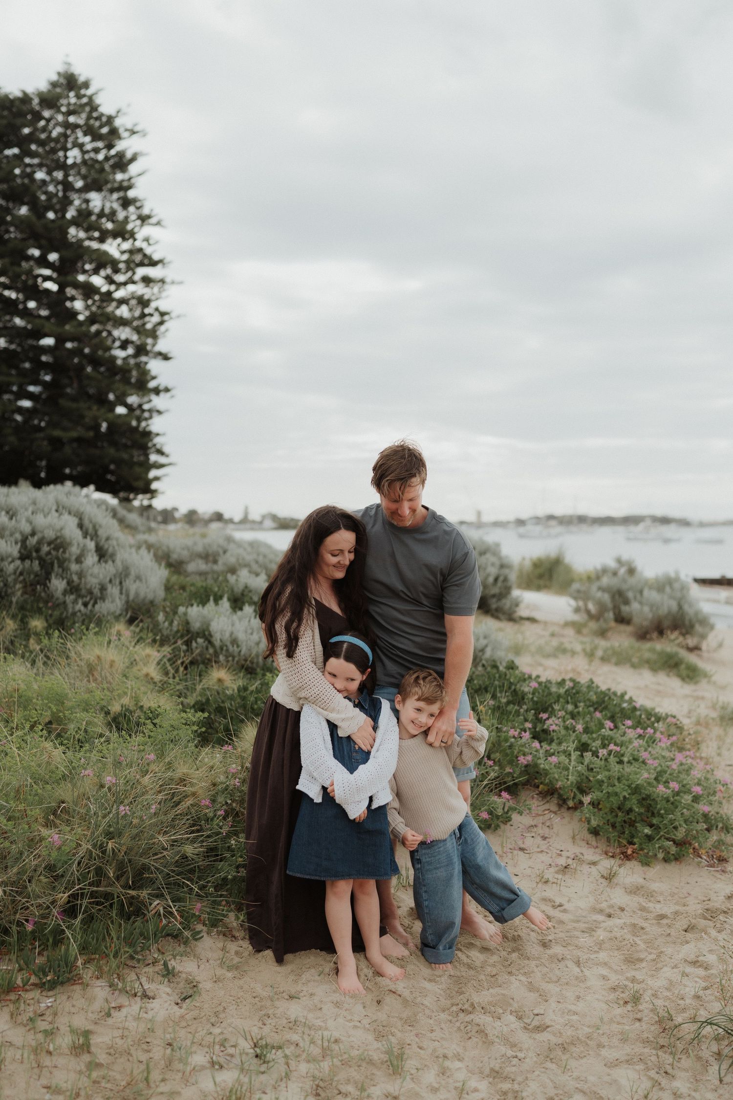 Family shares tender moment together while sitting on sandy beach dunes under cloudy sky.