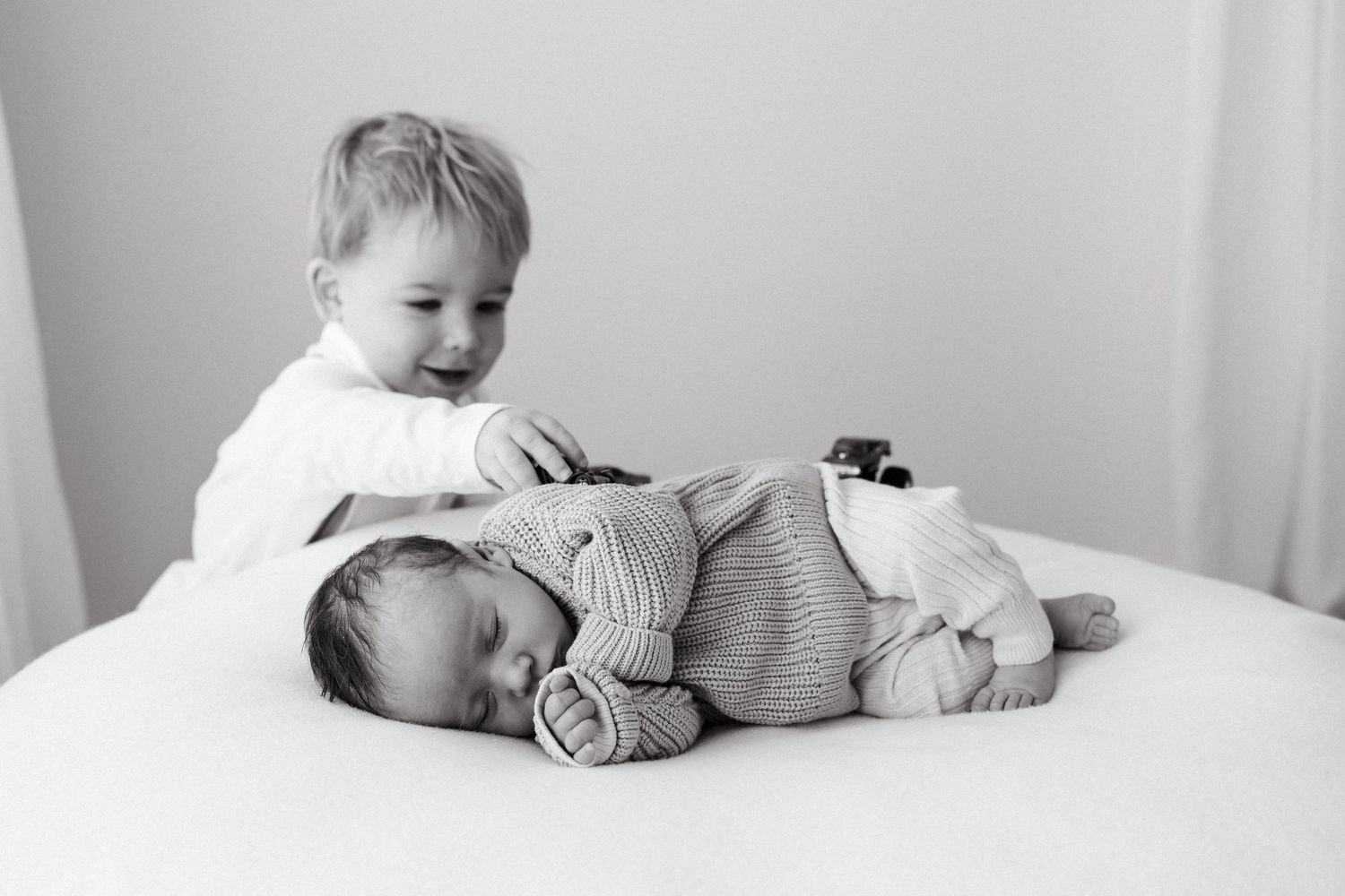 Two siblings share a tender moment as older sibling gently snuggles with sleeping baby on white bed.