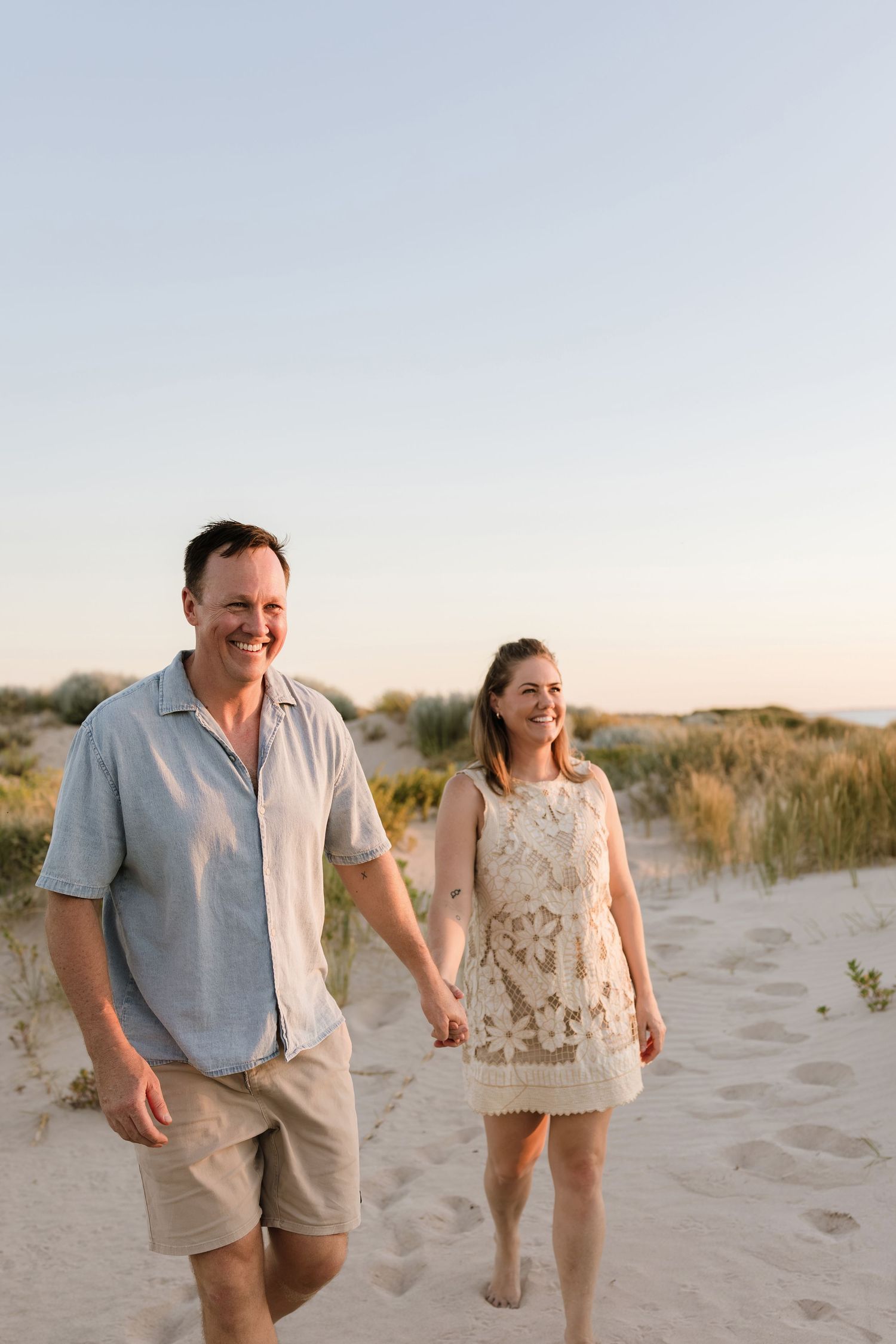 A couple walks hand in hand on a sandy beach at sunset wearing casual summer attire.