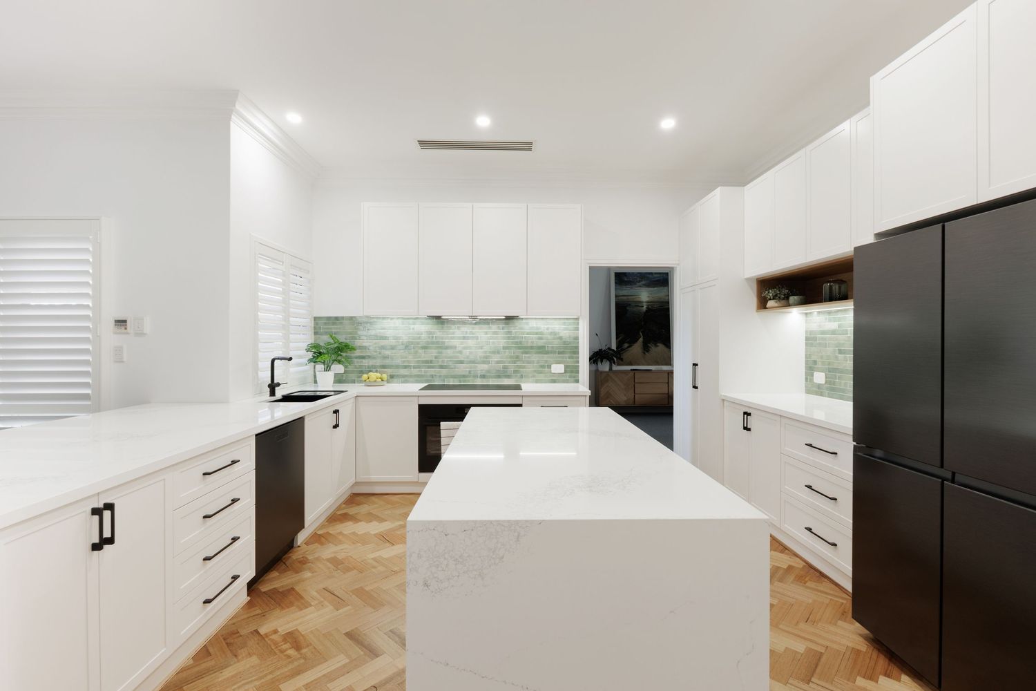 Modern white kitchen with marble island, herringbone timber floors, green tile backsplash, and black appliances.